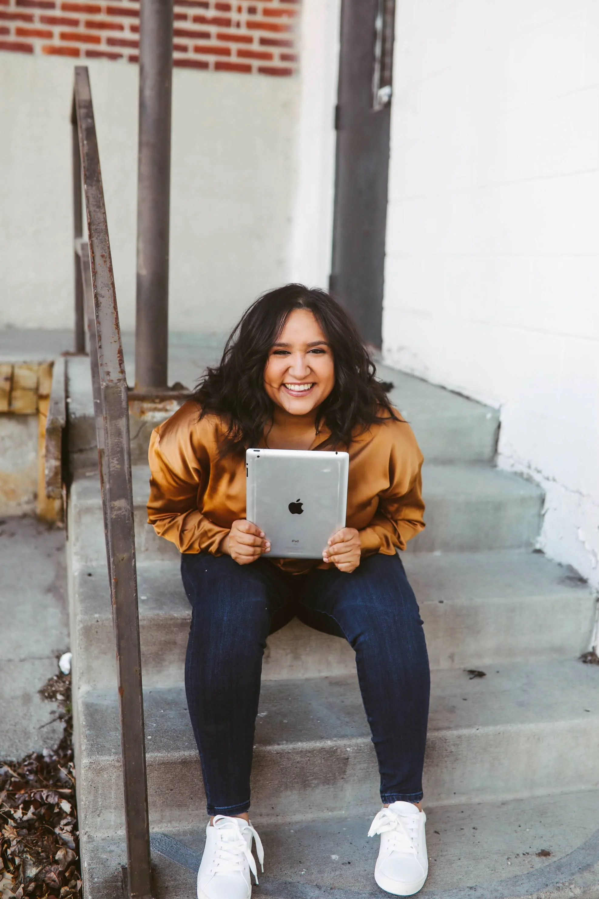 A woman sitting on outdoor steps, smiling and holding a tablet. She has long, dark curly hair and is wearing a bronze-colored jacket, blue jeans, and white sneakers. The background includes a brick wall and sidewalk.