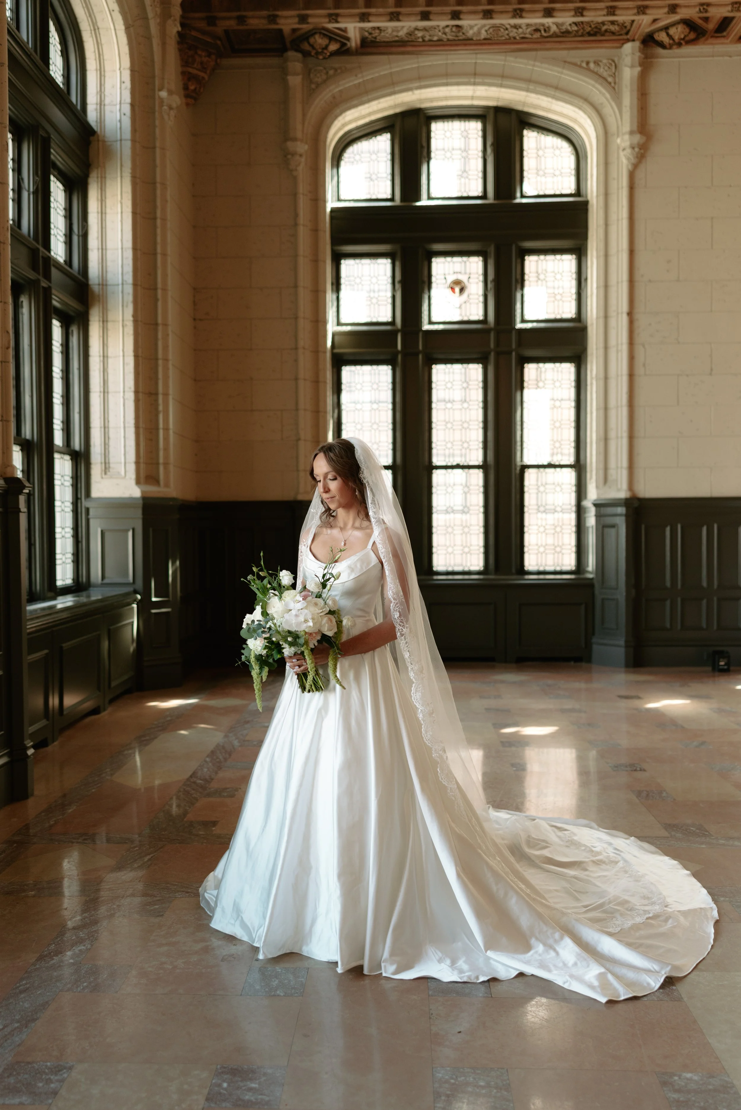 A bride in a white wedding gown with a lace veil, holding a bouquet of white and green flowers, standing in a large room with high ceilings and tall, stained-glass windows.