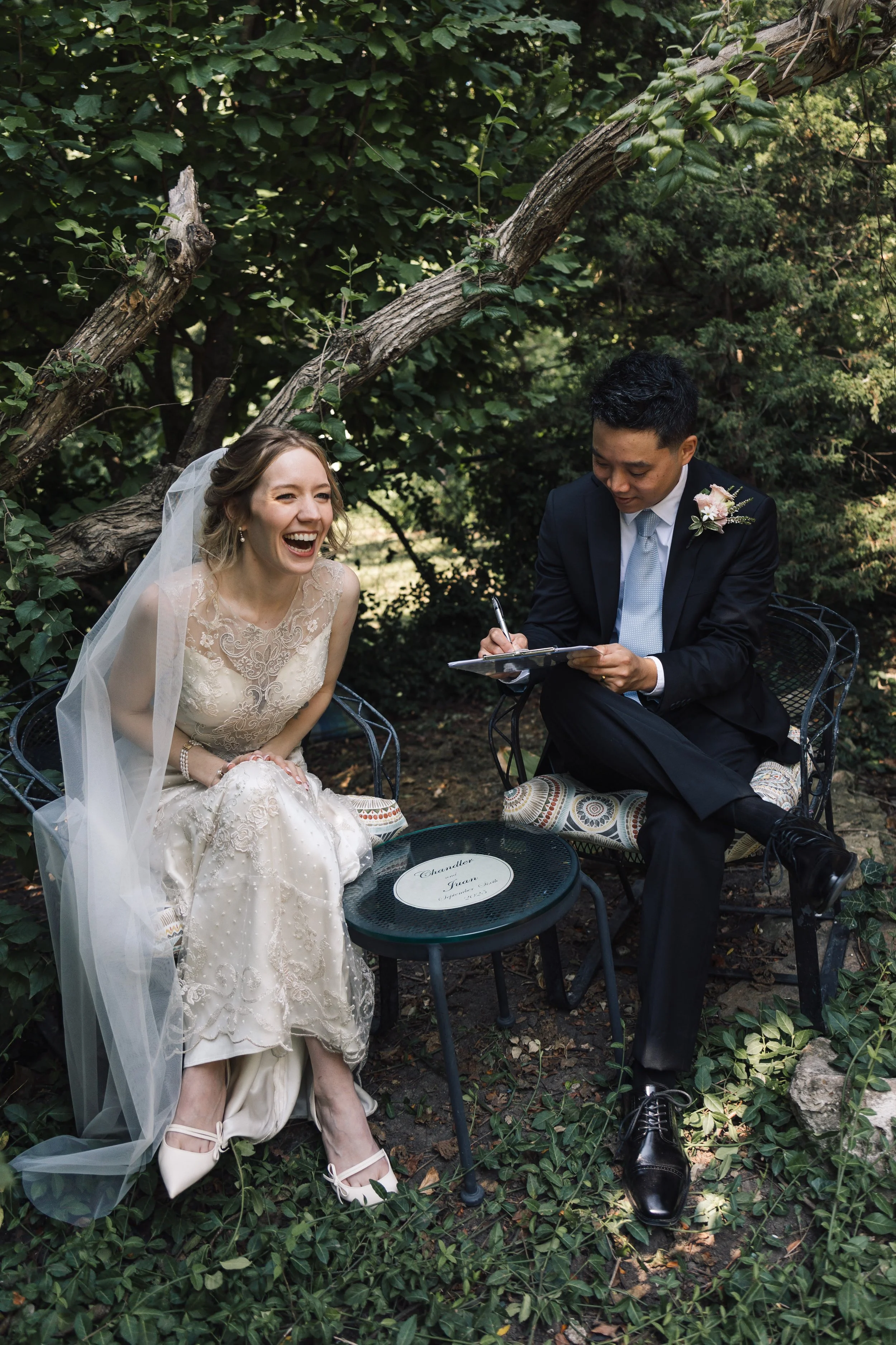 A bride and groom sitting outdoors among greenery, with the bride laughing and the groom signing a document.
