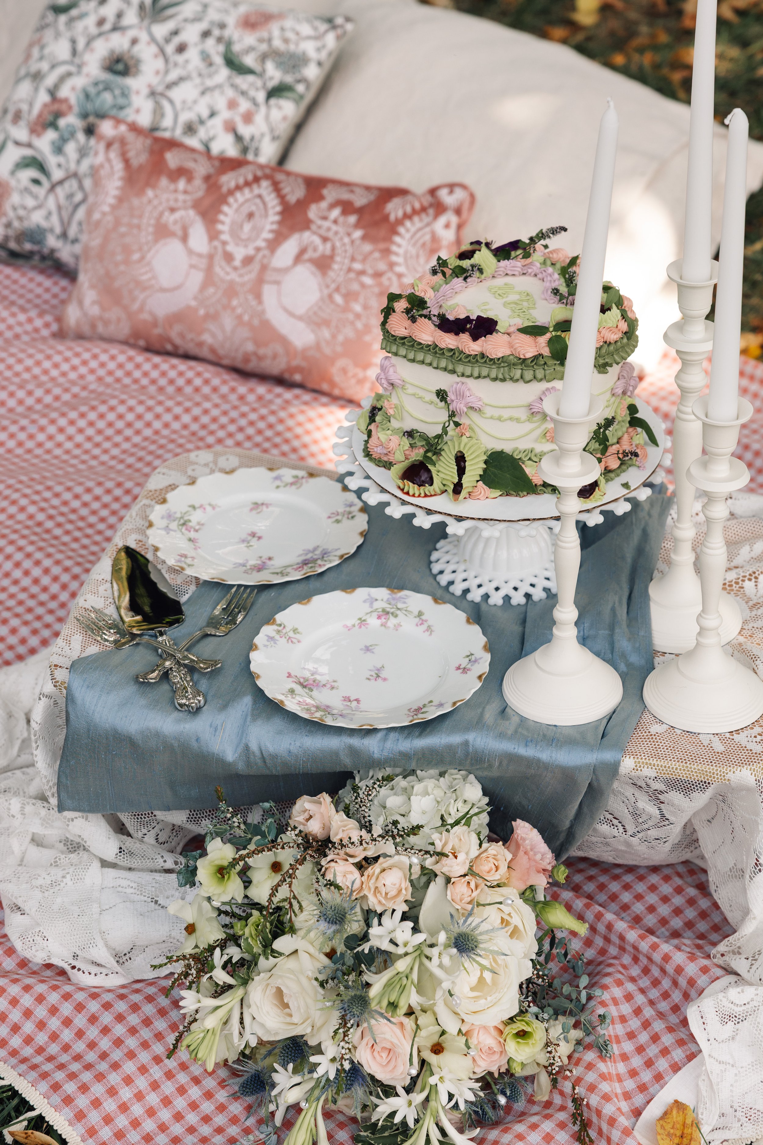 A decorated table with a floral wedding cake, floral arrangements, and vintage plates set on a red gingham tablecloth and lace fabric. Plush pillows are in the background.