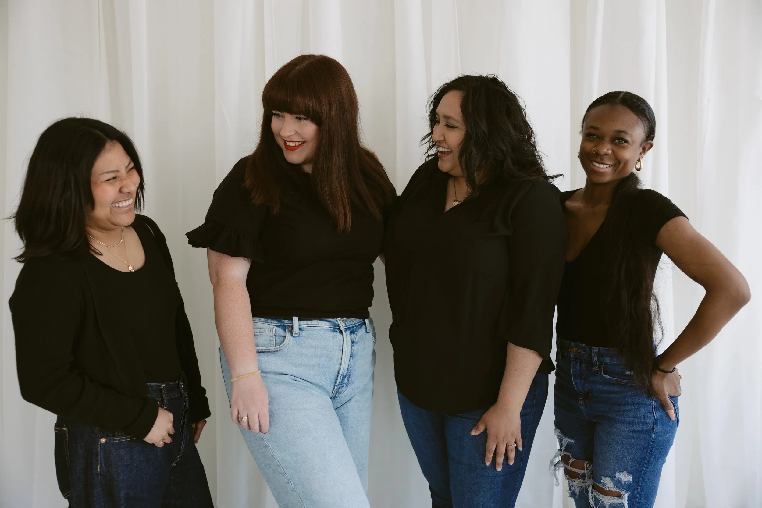 Four women standing together and smiling against a white curtain background, dressed in black tops and jeans.
