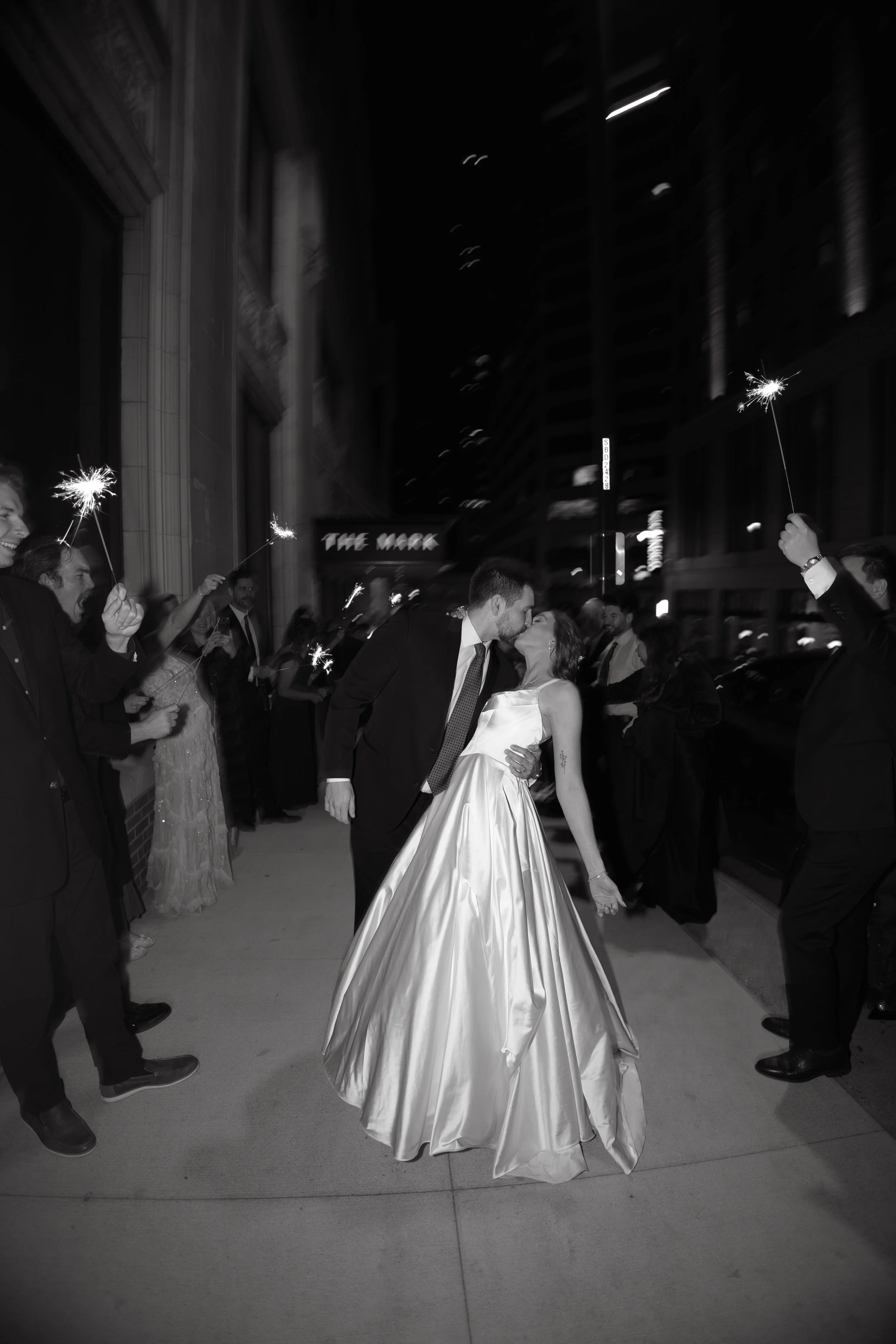 A couple in formal attire kissing on a city sidewalk at night, surrounded by people holding sparklers and celebrating.