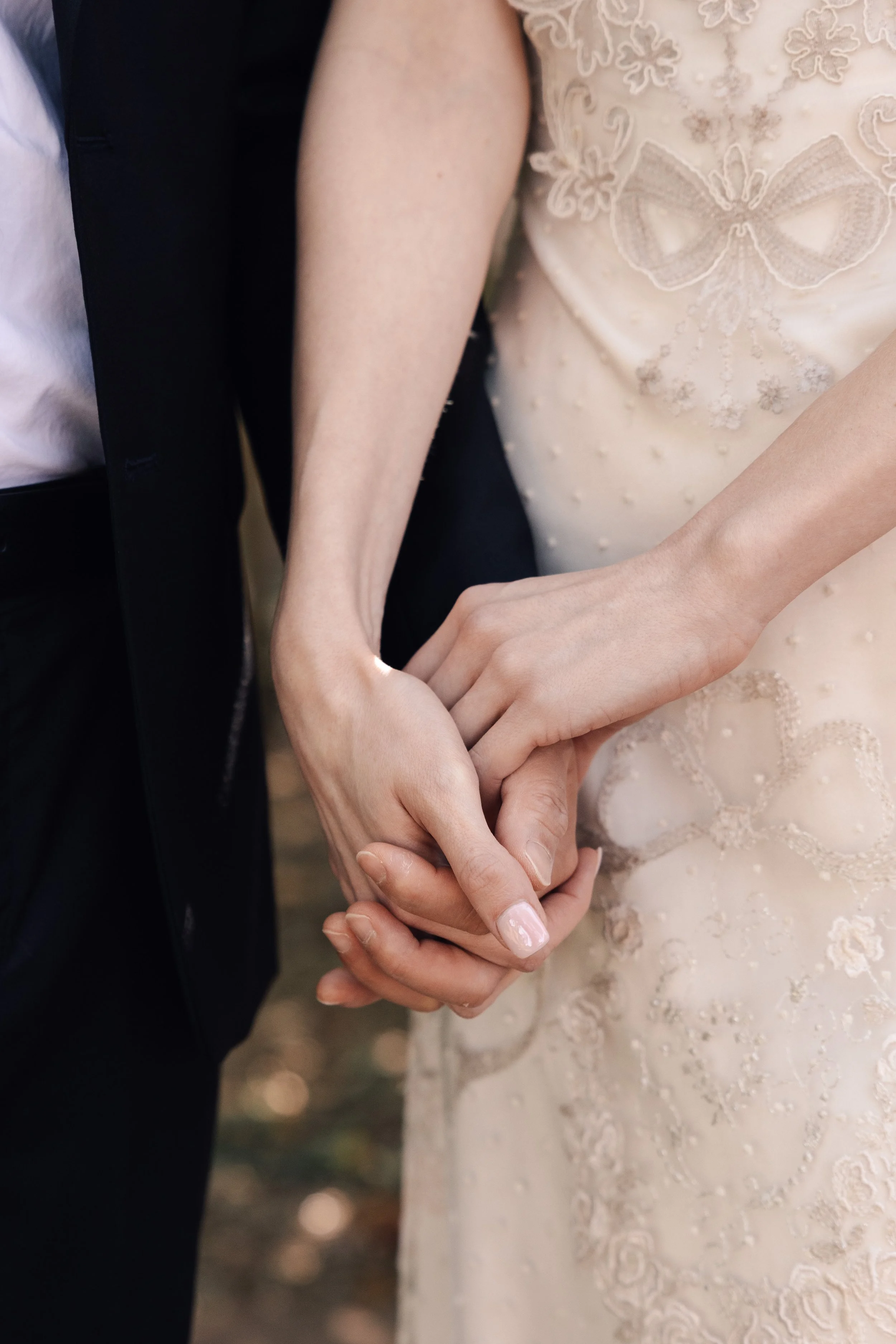 A close-up of two people holding hands, one in a black suit and the other in a beige dress with embroidery and a butterfly design.