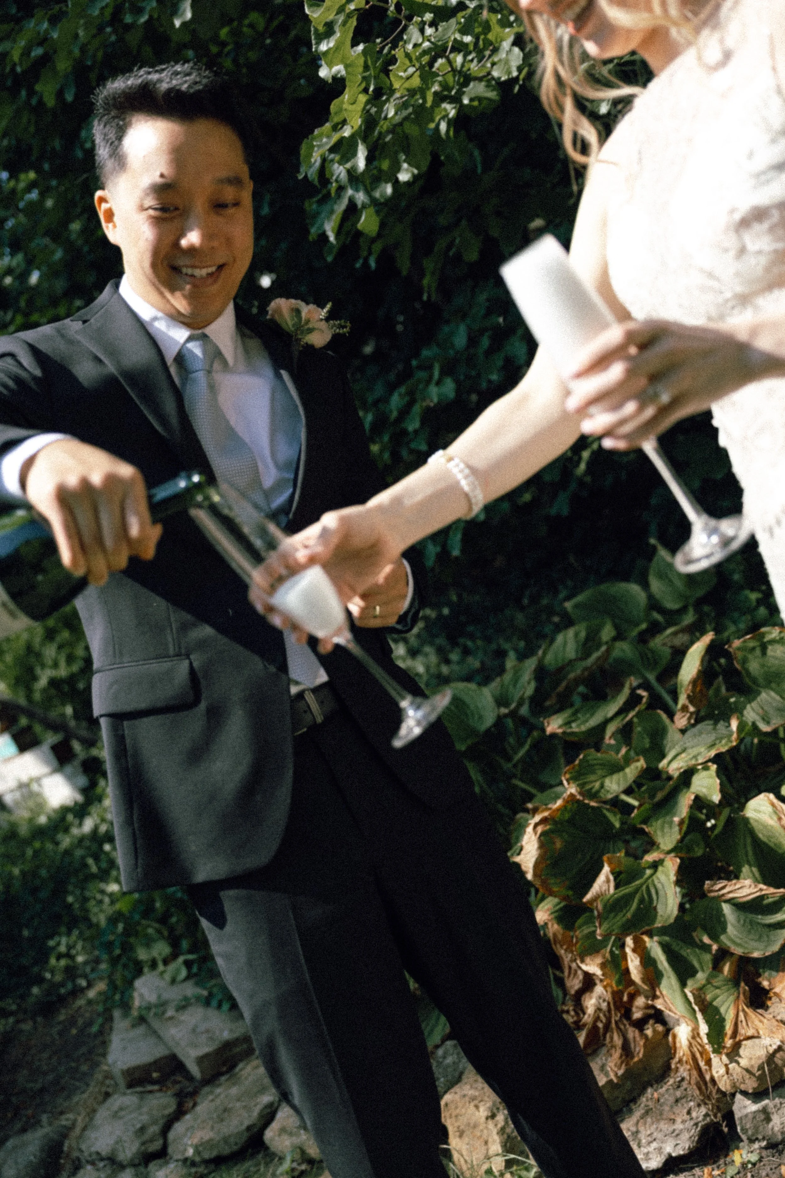 A man in a suit is pouring champagne into a woman's glass during a celebration at night outdoors, with greenery in the background.