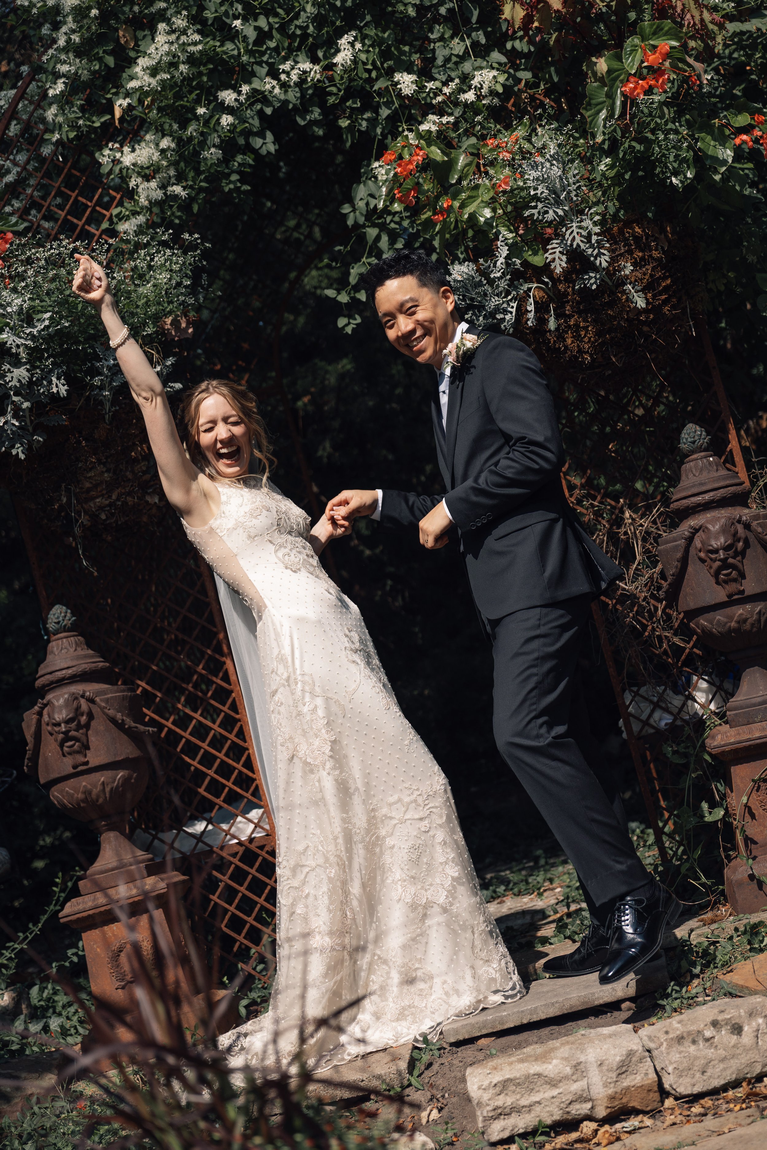 A smiling bride and groom celebrating outdoors under a floral arch, holding hands, with lush greenery and flowers in the background.
