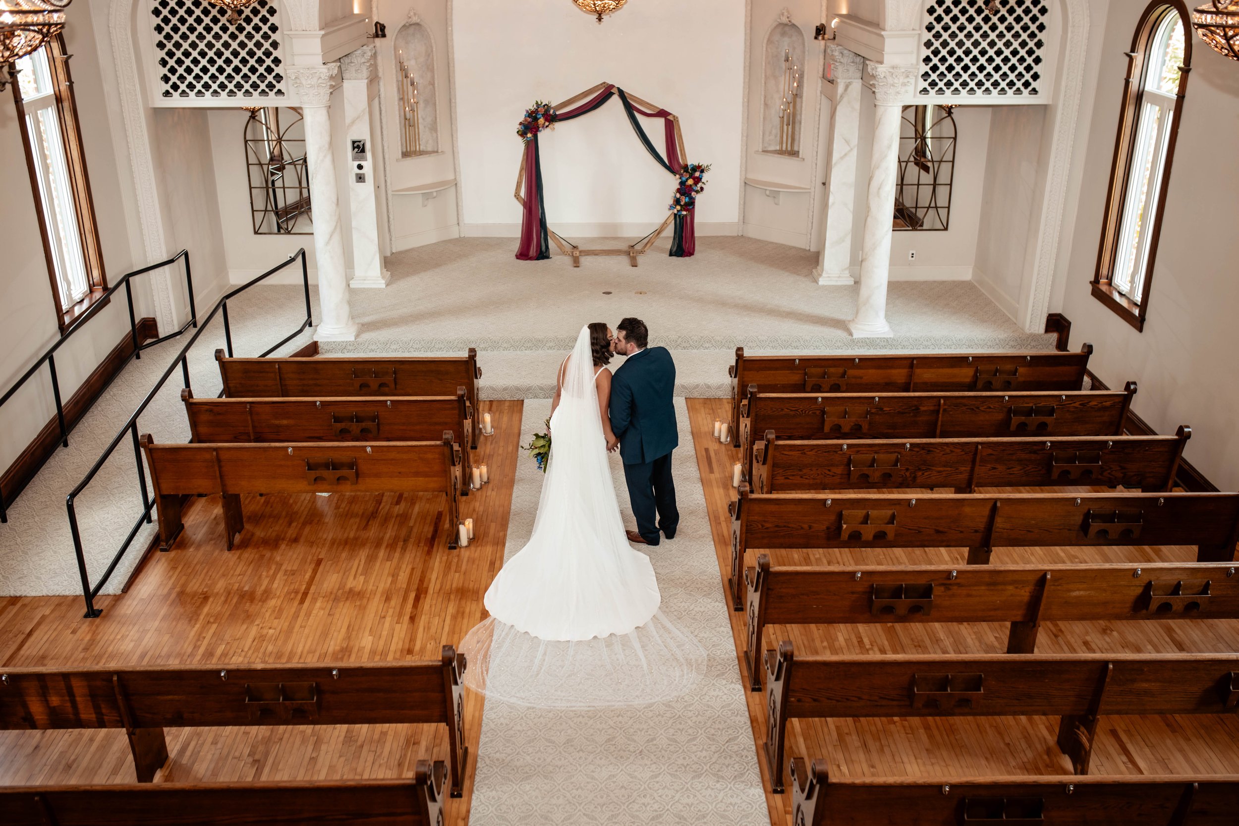 A bride and groom standing and kissing at the altar in a wedding ceremony, in a small chapel with wood pews, columns, and floral decorations on the frame of the altar.