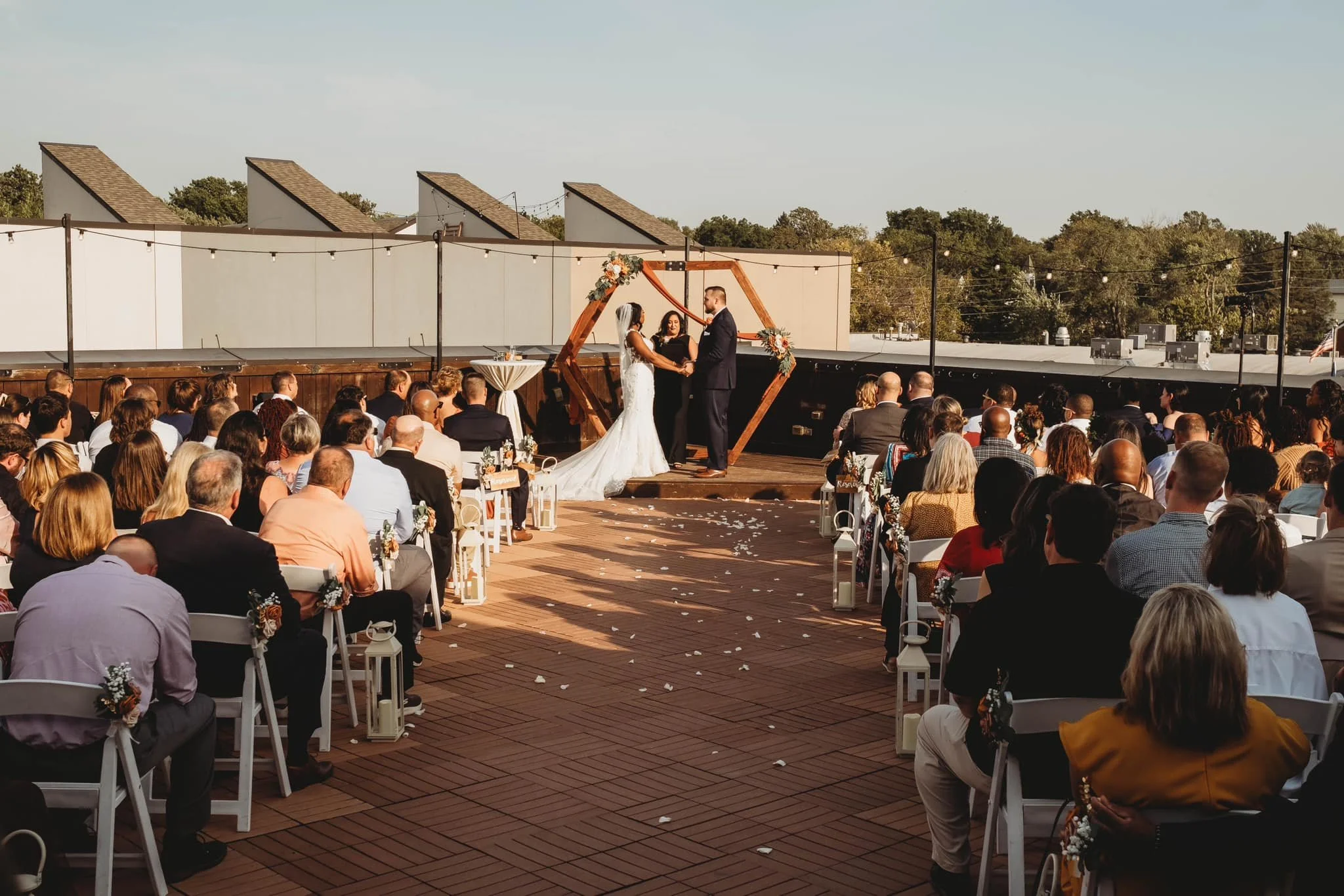 Outdoor wedding ceremony with bride and groom holding hands, officiant standing between them, guests seated on both sides, decorated arch and string lights, rooftop setting amidst trees and rooftops.