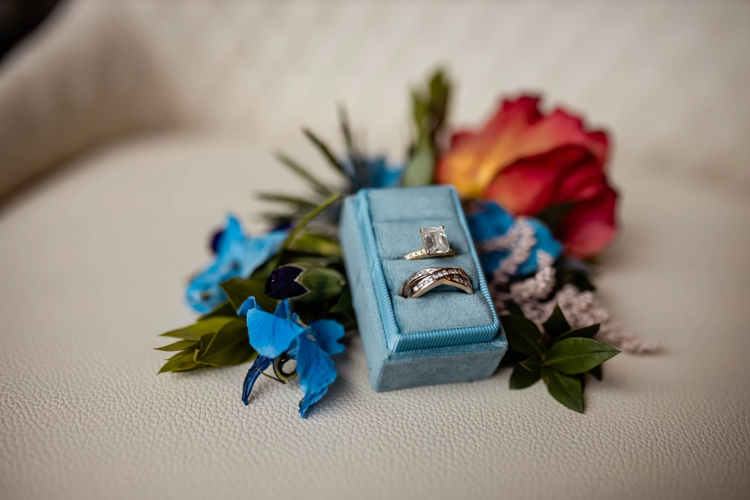 A jewelry ring with a large rectangular gemstone displayed in a blue box, surrounded by a colorful floral arrangement, on a light textured surface.