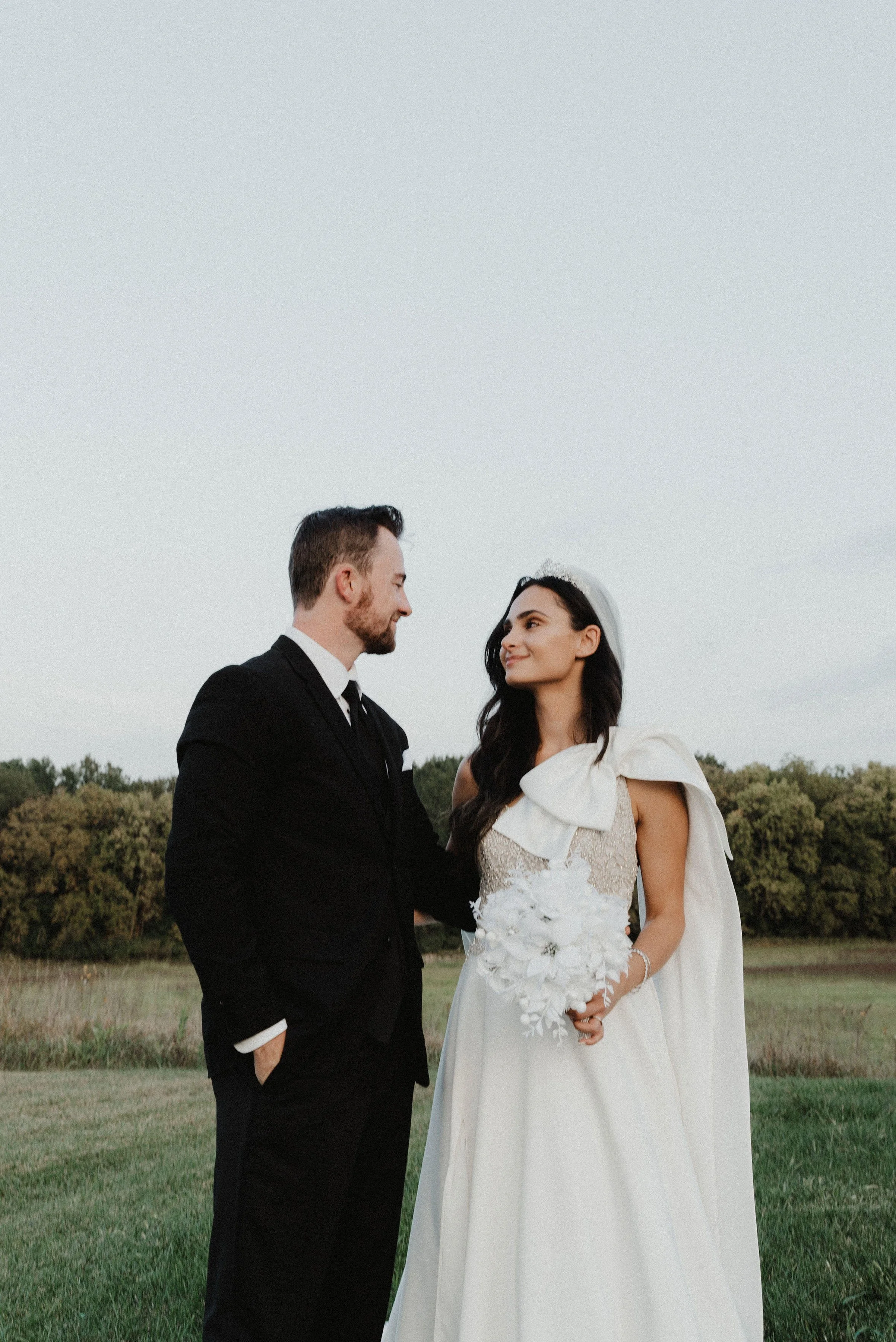 A couple dressed in wedding attire standing outdoors in a grassy area with trees, gazing into each other's eyes during sunset.
