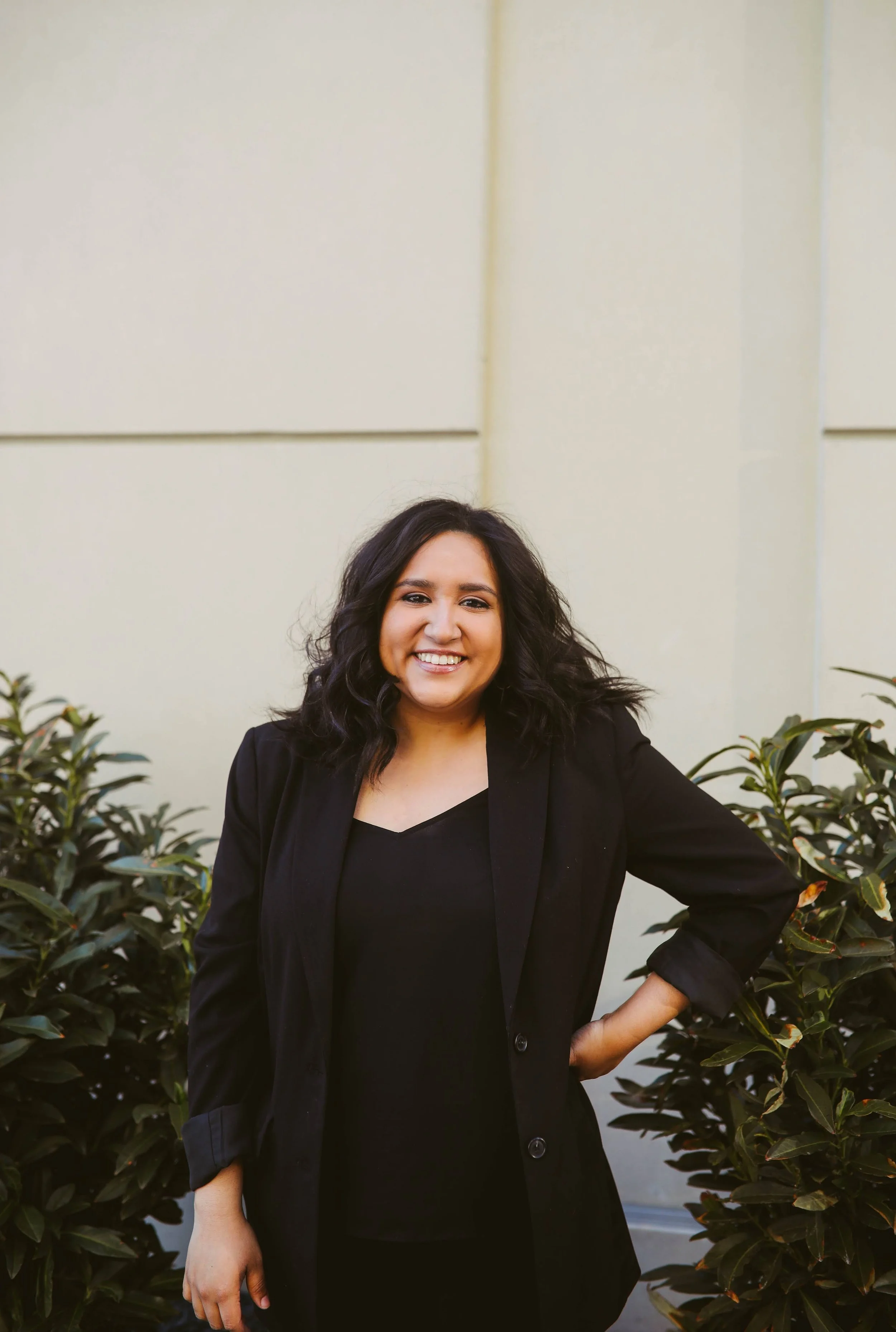 A woman with dark, wavy hair smiling and wearing a black blazer, standing outdoors against a plain, light-colored wall with green plants on either side.
