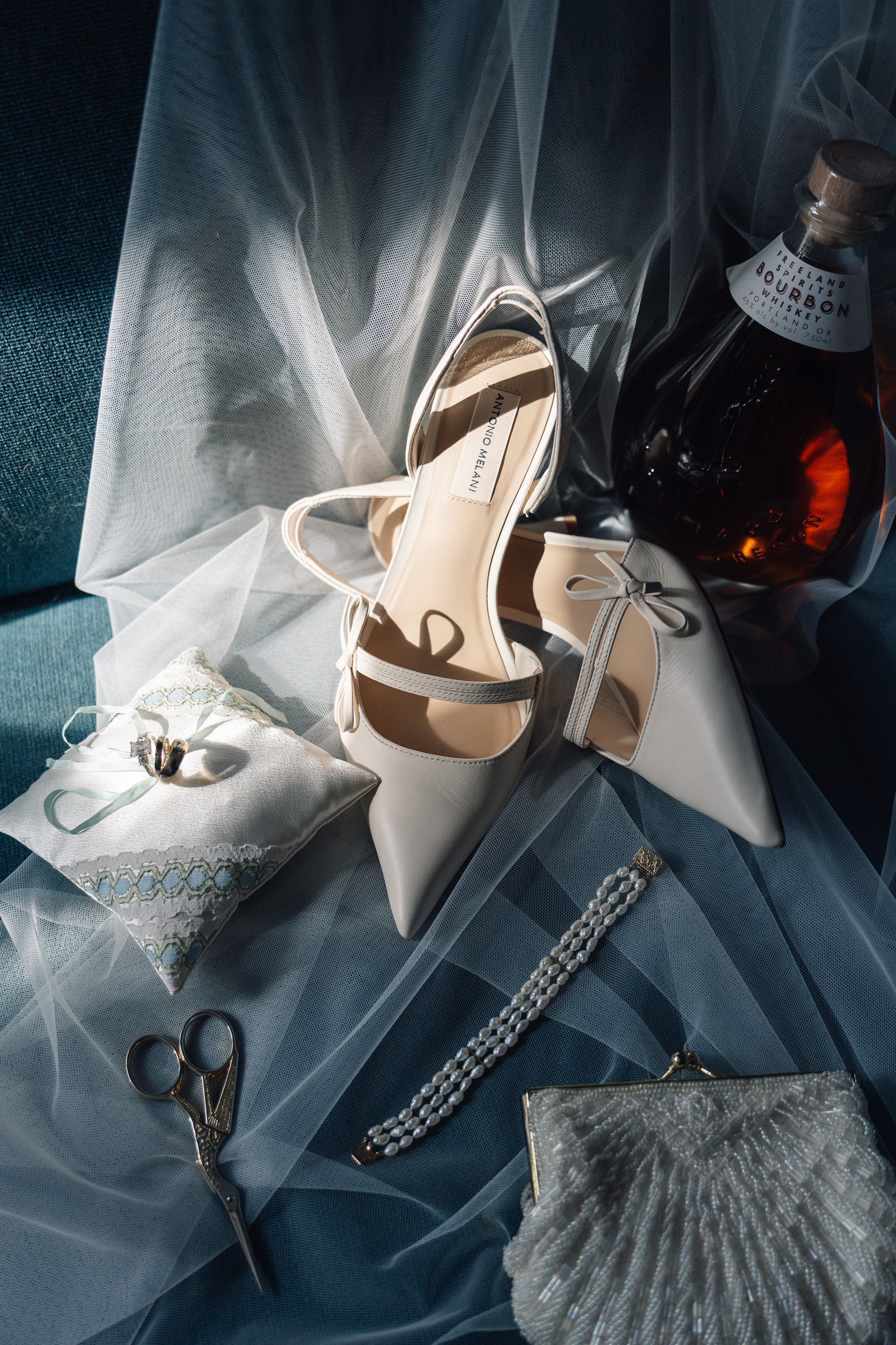 Elegant flat-lay of wedding accessories on a dark blue fabric surface, including a pair of white Antonio Melani high heels, a pearl bracelet, a pair of wedding rings, a small decorative pillow, a clutch purse, and a bottle of whiskey with a black and