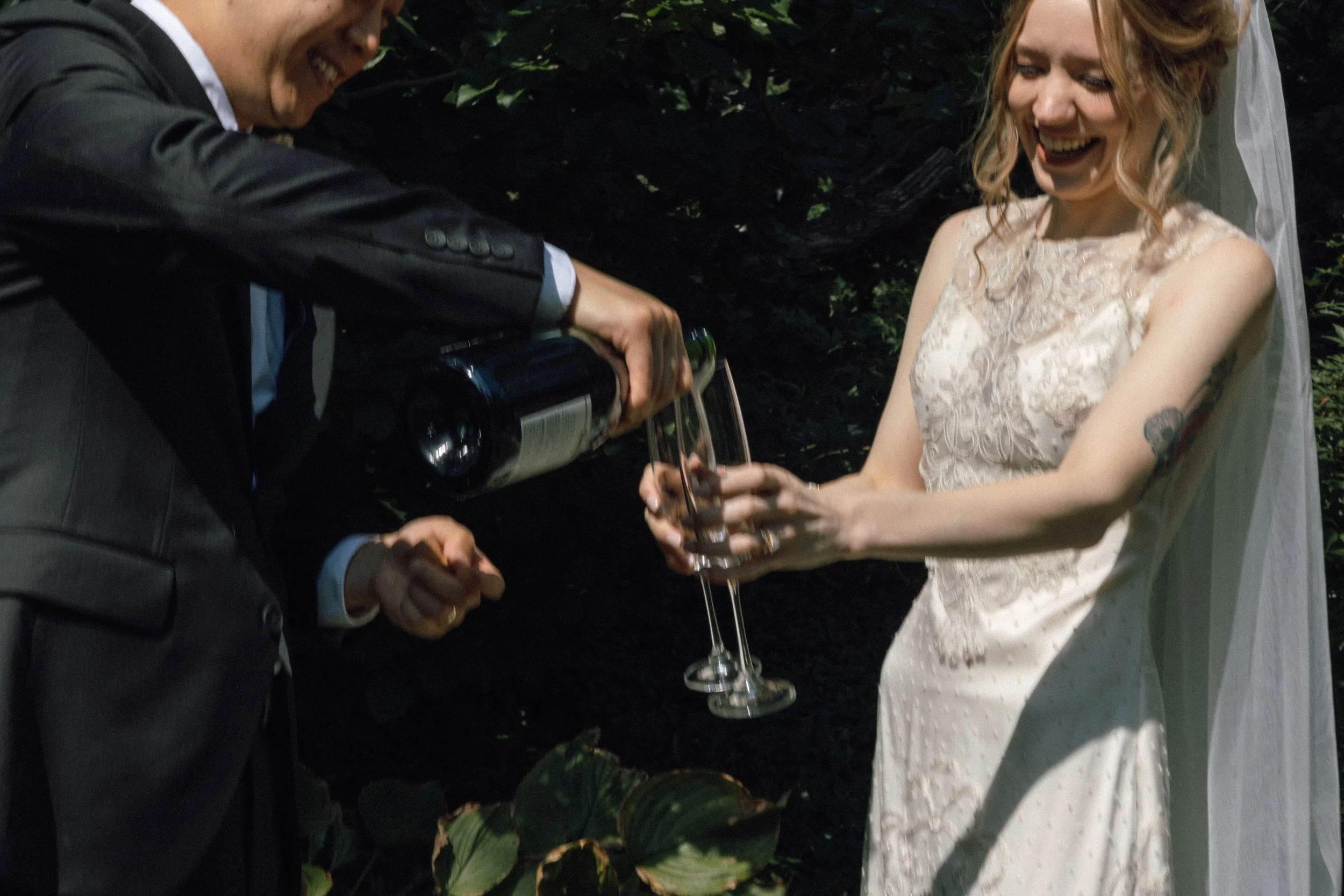 A man and a woman dressed in wedding attire sharing a toast with champagne outdoors surrounded by greenery.