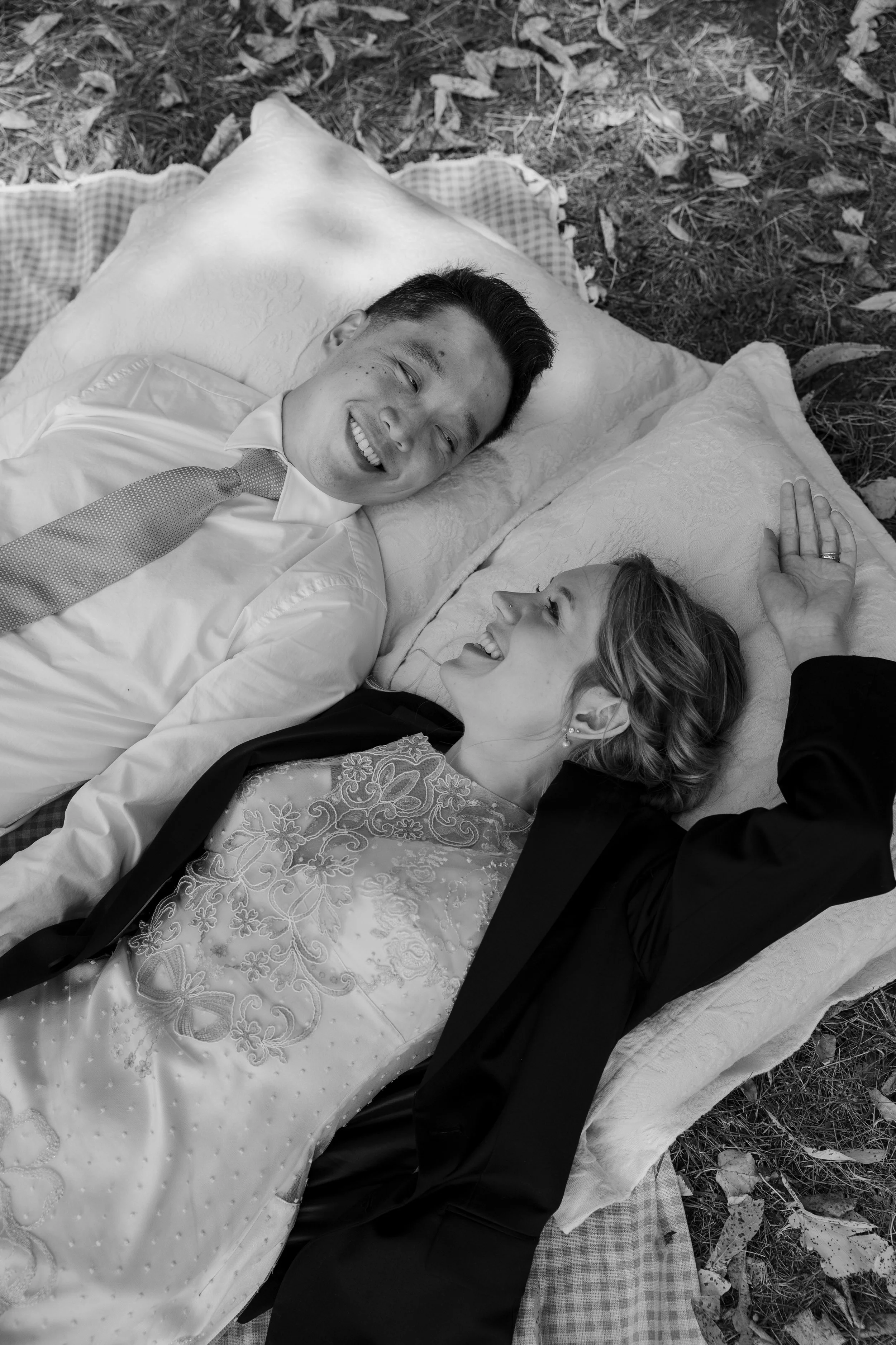 A smiling man and woman lie on a blanket outside, looking at each other, surrounded by fallen leaves.