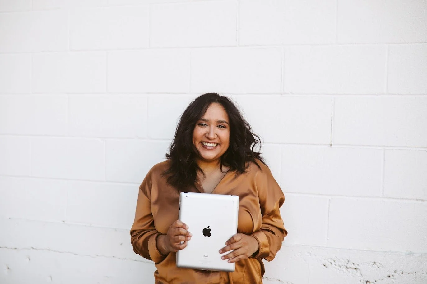 A smiling woman with dark wavy hair holding an Apple iPad in front of a white brick wall.