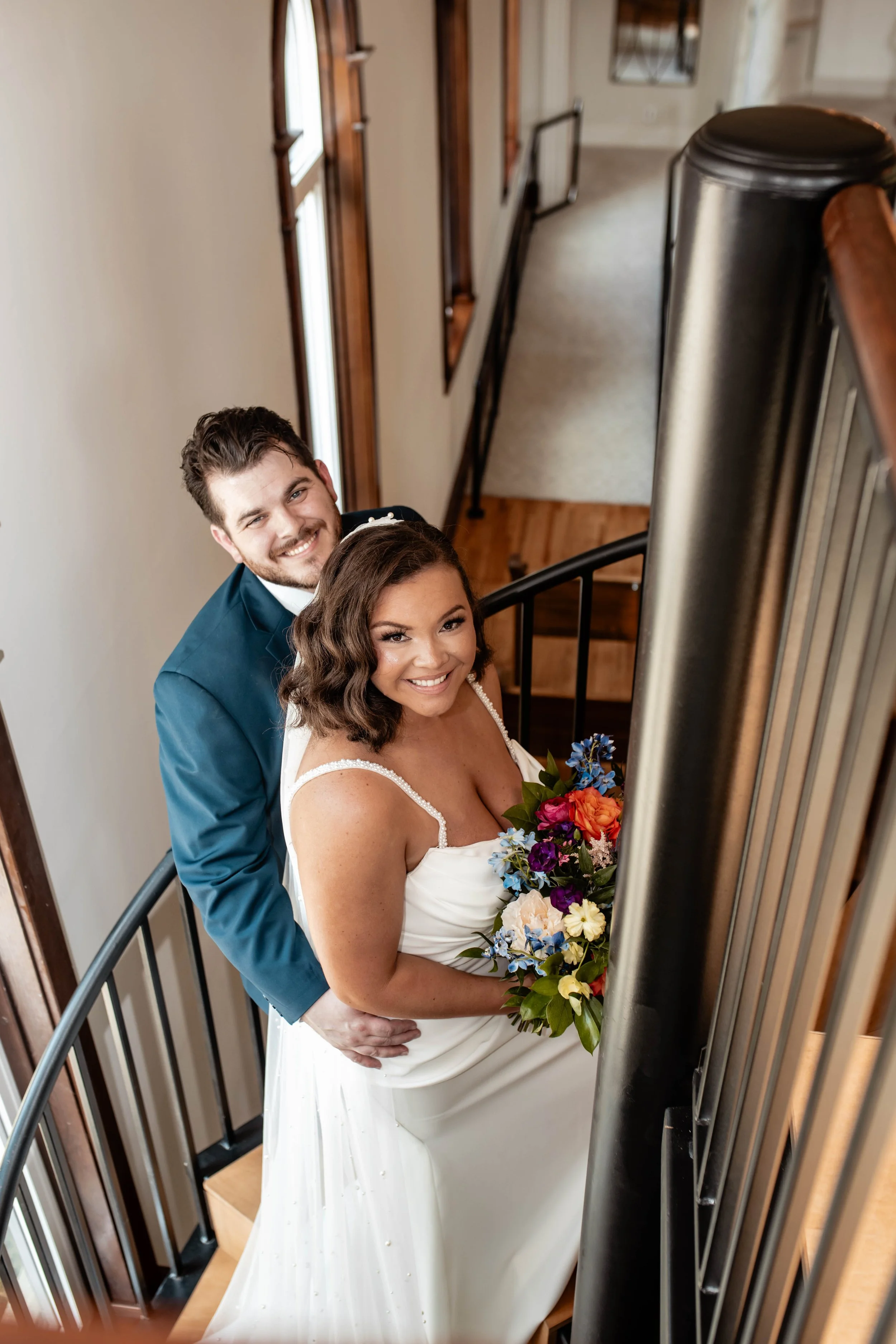 A bride and groom on a staircase, smiling and looking up at the camera. The bride is holding a bouquet of colorful flowers, and the groom is dressed in a blue suit. The staircase has black railings and wooden accents, with windows and wall decor visi