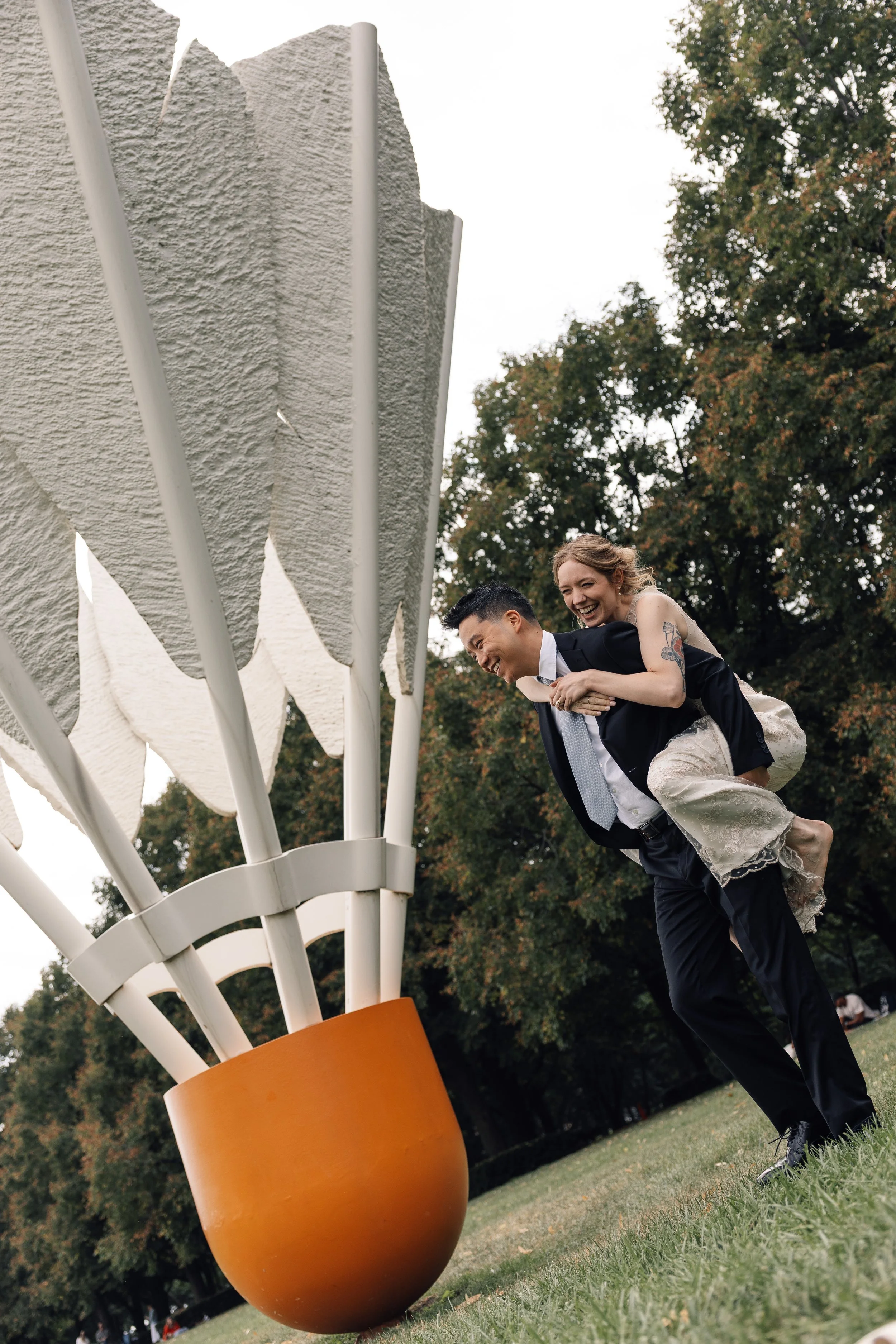 A couple riding a bicycle tandem, with the man in a black suit and the woman in a wedding dress, outdoors in a park with trees, near a large orange flower sculpture.