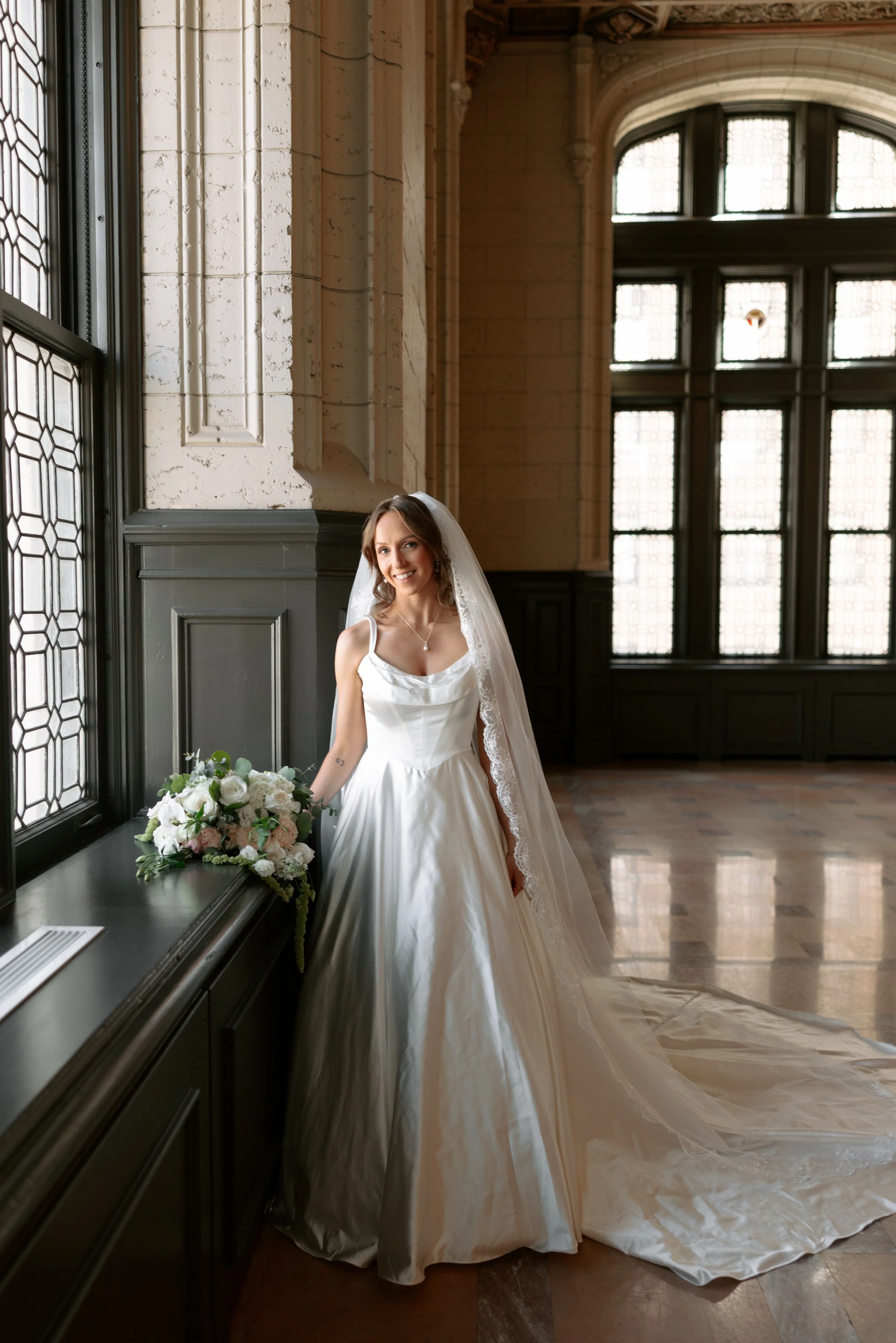 A smiling bride in a white wedding gown standing indoors near large windows and a flower arrangement, with a long train of the dress flowing on the floor.
