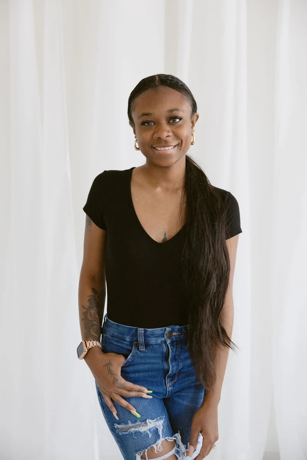 A young African American woman with long black hair tied to the side, wearing a black V-neck shirt and ripped blue jeans, standing in front of white curtains, smiling at the camera.