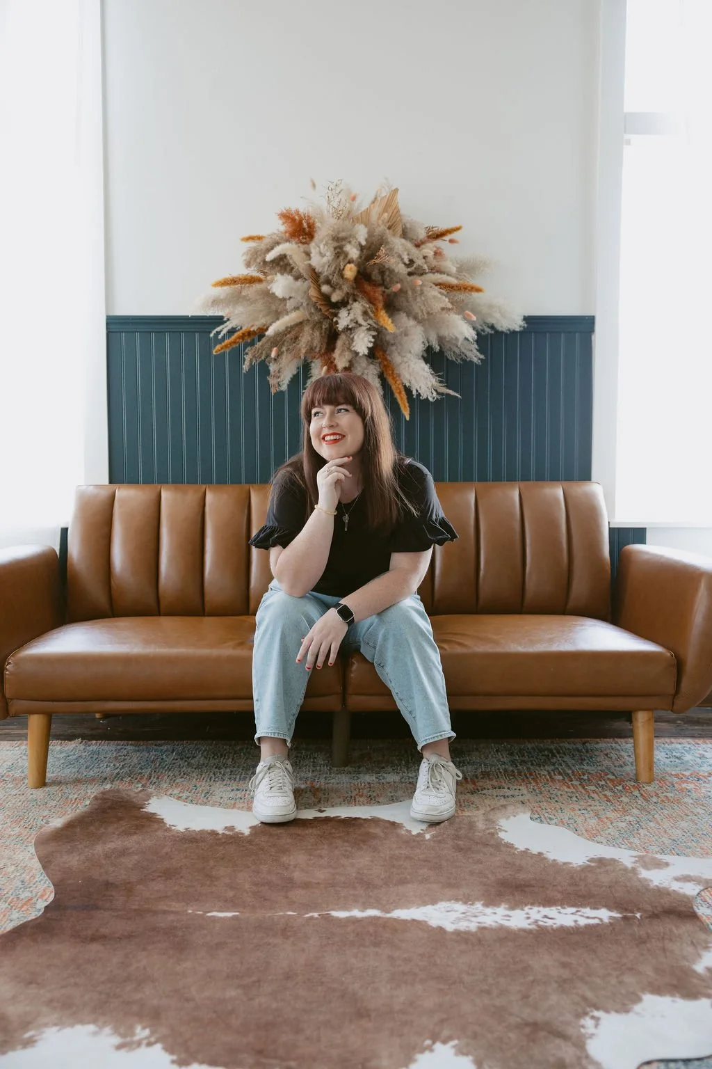 A woman sitting on a tan leather sofa with a decorative dried flower arrangement above her head in a modern, well-lit room.