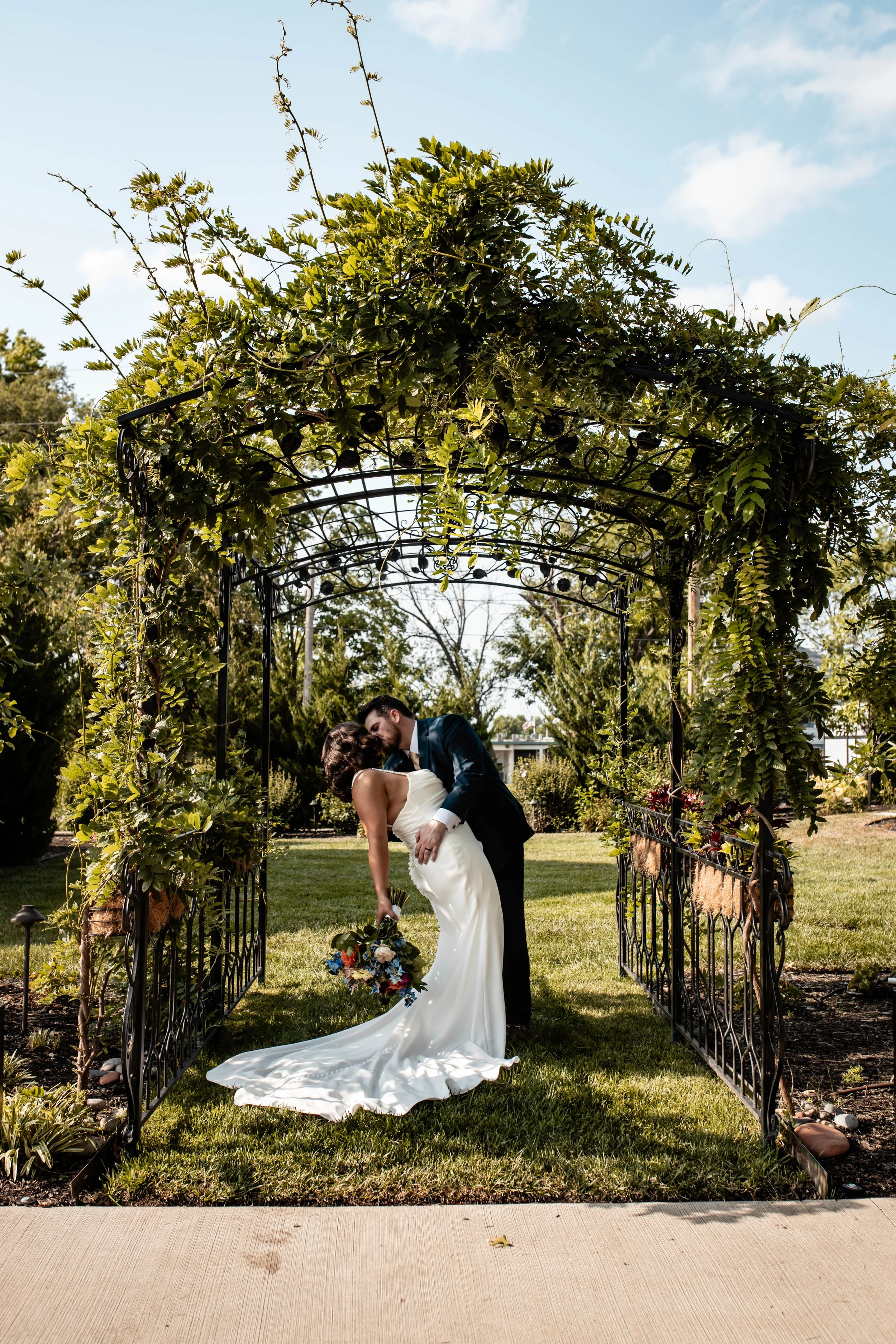 A bride and groom sharing a kiss under a garden arch decorated with green foliage and vines, the bride holding a colorful bouquet of flowers, outdoors on a sunny day with trees in the background.