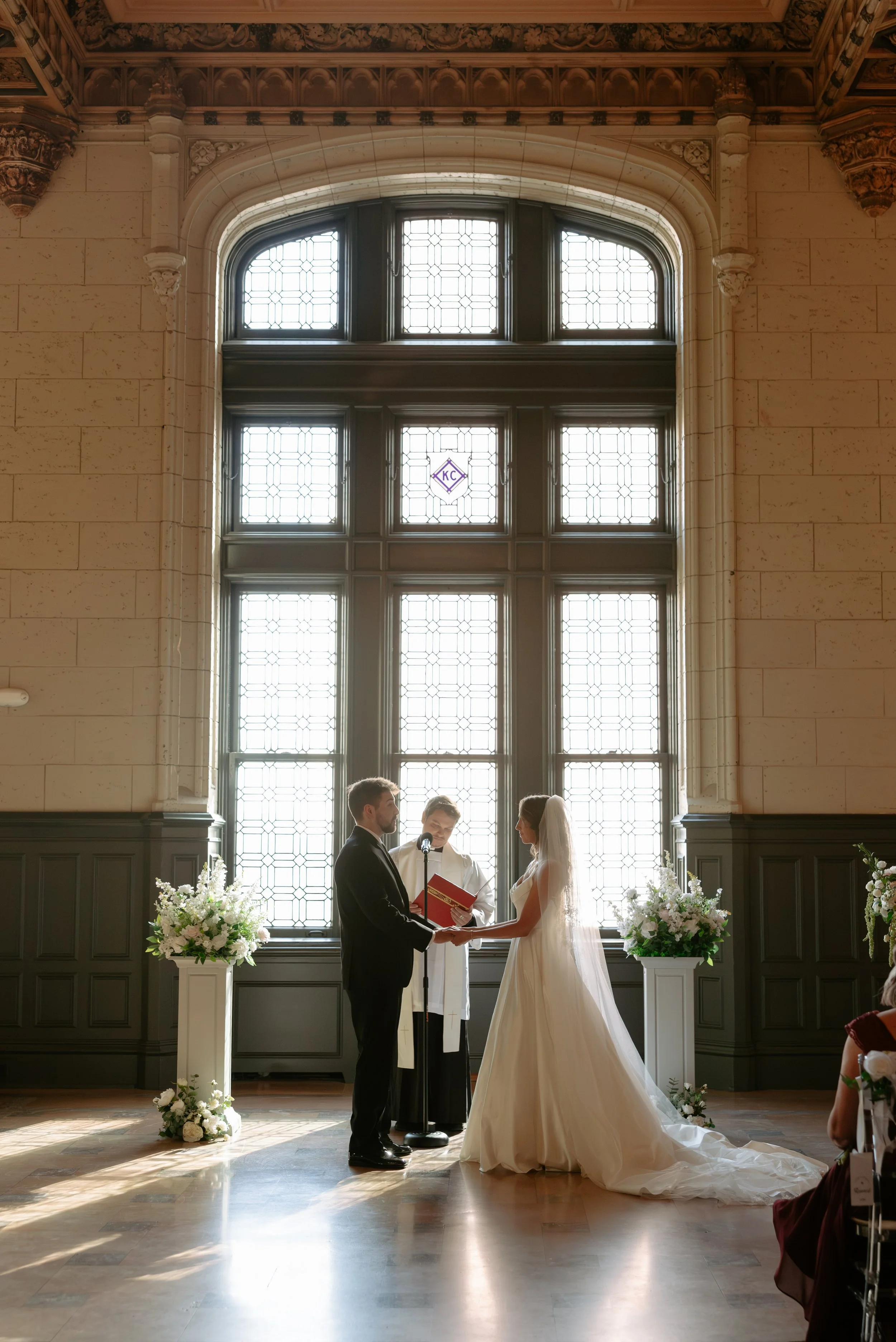 A wedding ceremony taking place indoors with a bride and groom facing each other, holding hands, with an officiant between them. Large stained glass window in the background with sunlight streaming through, decorated with floral arrangements.