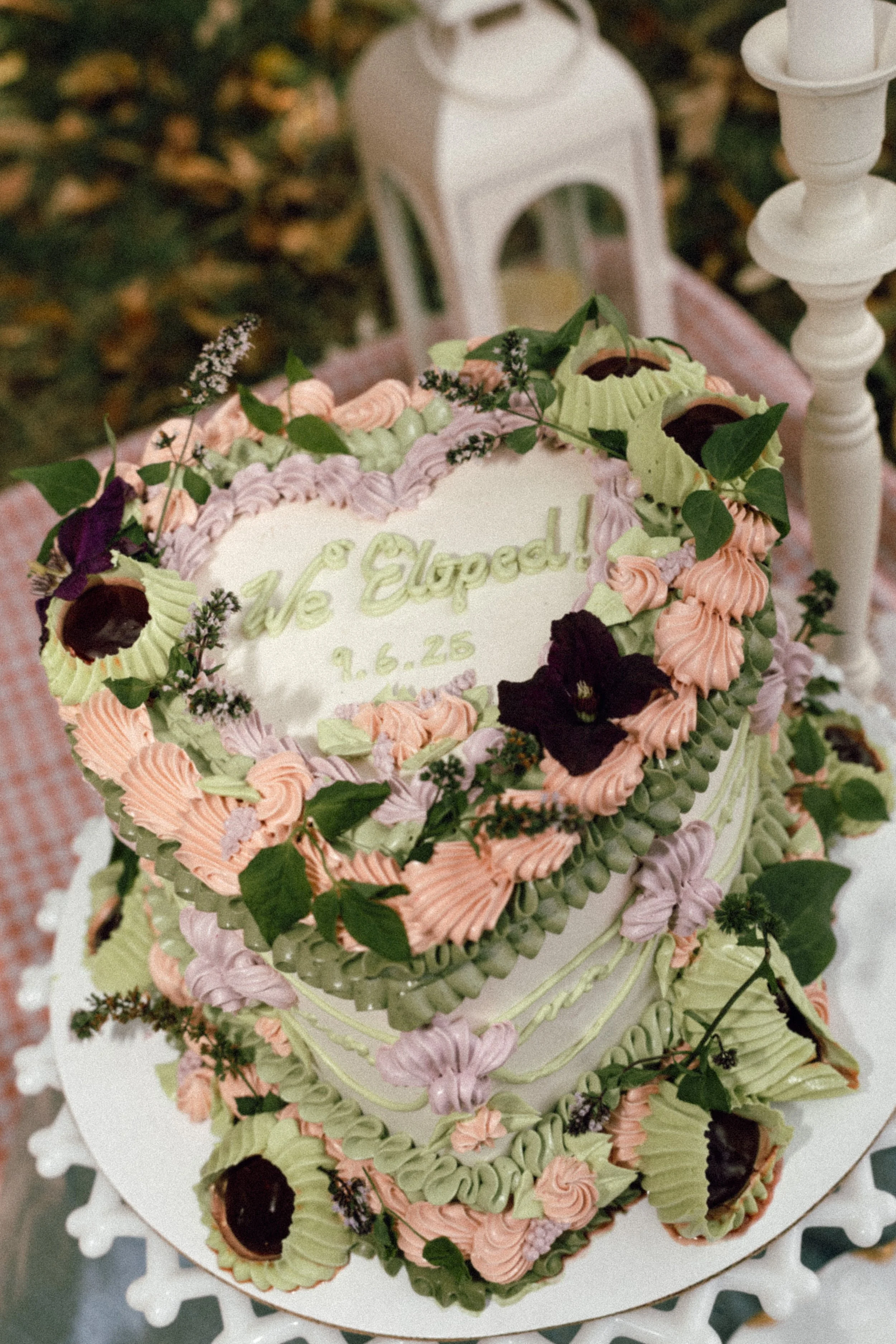 A decorative cake with pastel-colored icing, flowers, and greenery on top, placed on a white cake stand with ornate edges. The cake has a message written in green icing that reads 'We Expected' and a date '9.6.25' visible at the center.