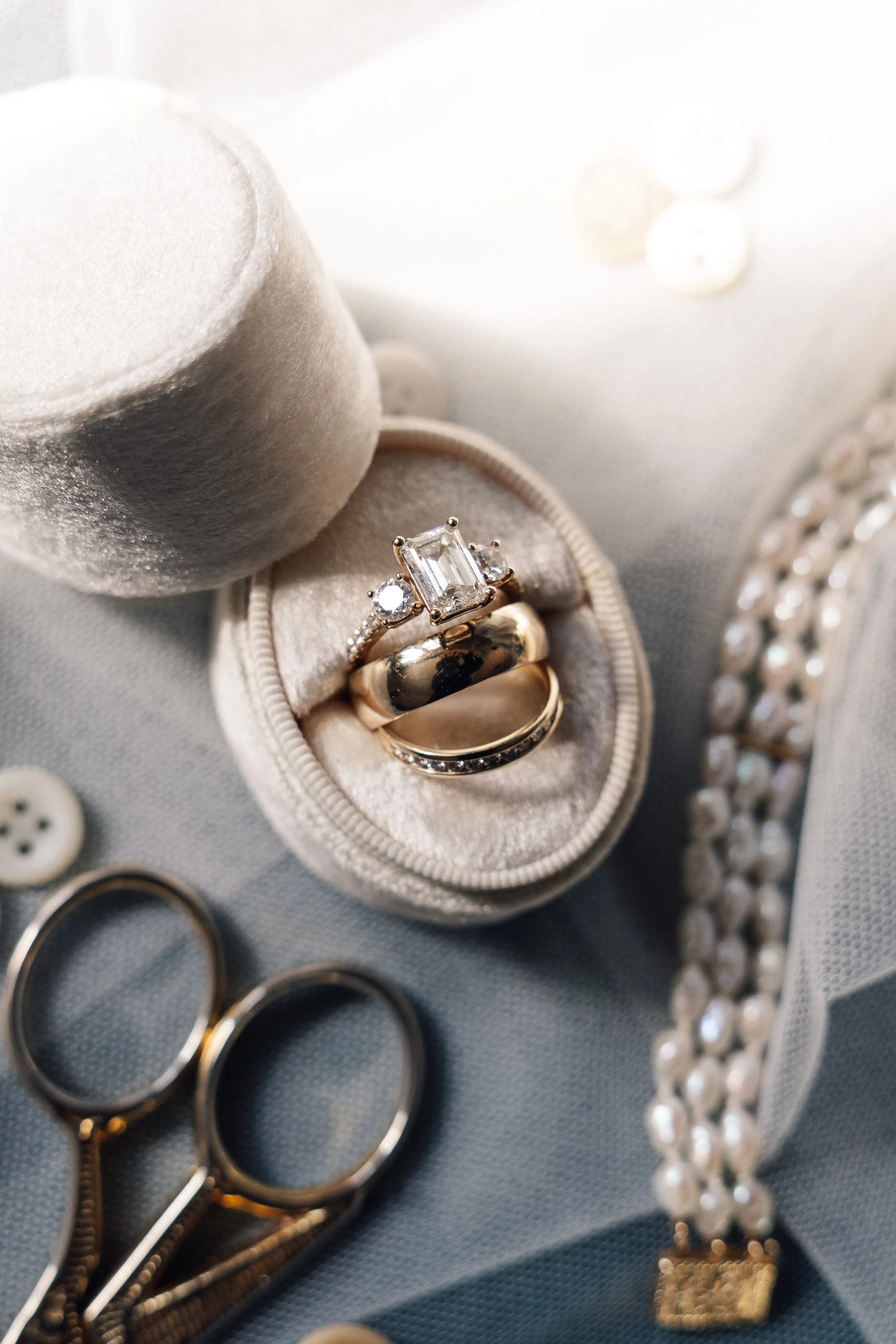 Close-up of rings in a jewelry box surrounded by pearls, scissors, buttons, and small decorative items on fabric.