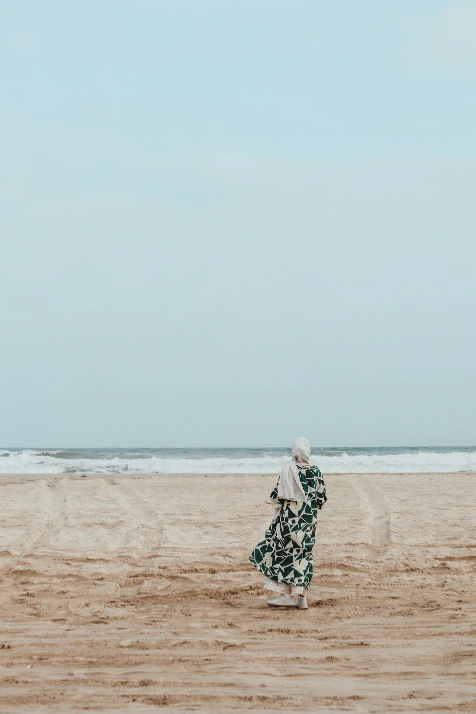 A person wearing a hijab and a patterned dress walking on a sandy beach facing the ocean with waves and a cloudy sky.