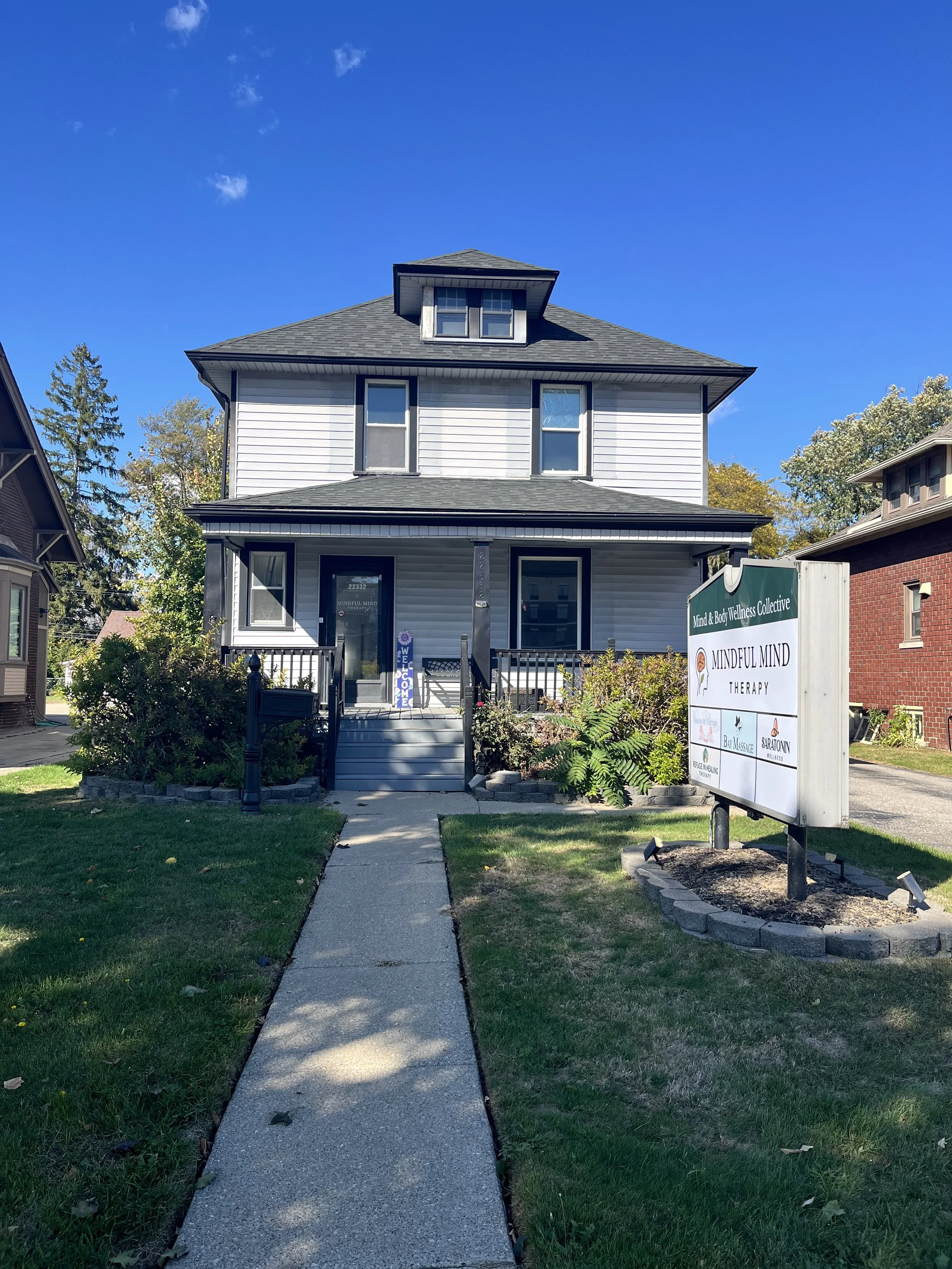 A two-story house with white siding and black trim, with a front porch and a sidewalk leading to the entrance. The house has a dormer window on the roof and is surrounded by a lawn with bushes and trees. There is a sign in the yard that reads "Mind & Body Wellness Collective" and "Mindful Mind Therapy".
