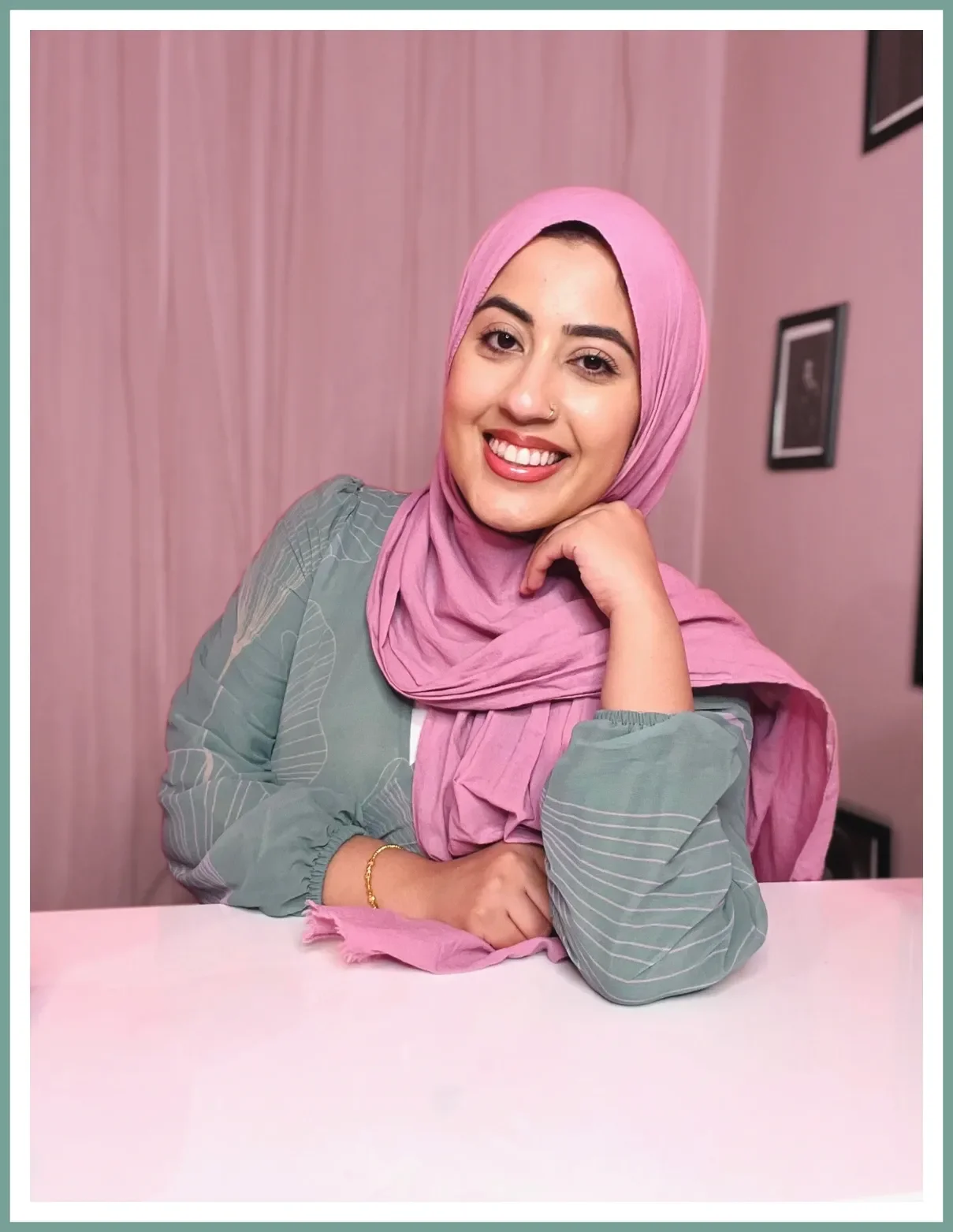 A young woman wearing a pink hijab and a gray top sitting at a pink table, smiling, with a pink and gray background with framed pictures.