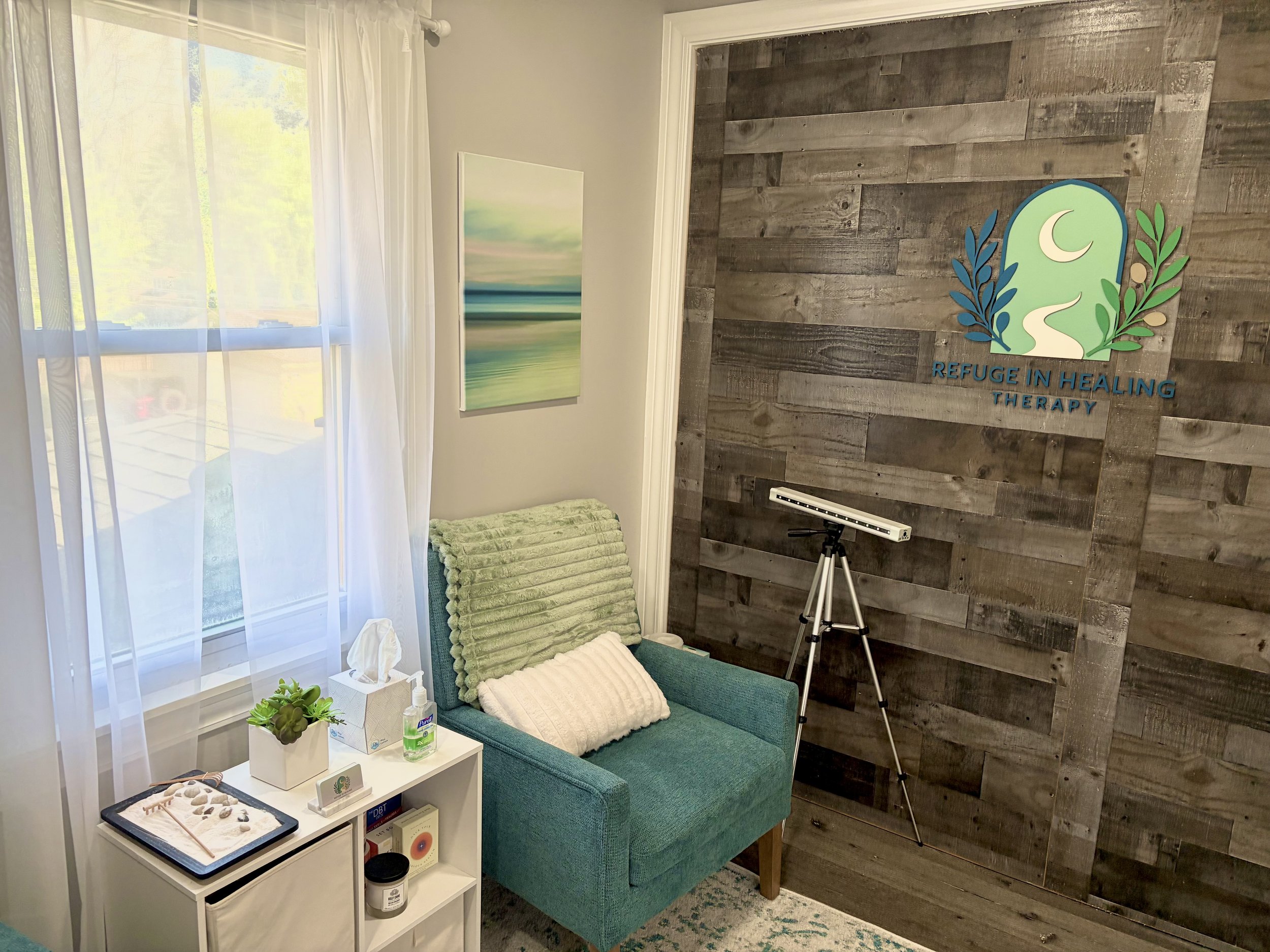A cozy therapy room with a teal couch, a white side table with a plant, tissues, hand sanitizer, and informational cards, a wall-mounted art piece of blurred landscape, and a dark wood accent wall with a logo that reads "Refuge in Healing Therapy" featuring a crescent moon, a winding path, and leafy branches. A tripod with a device is also present in the room.