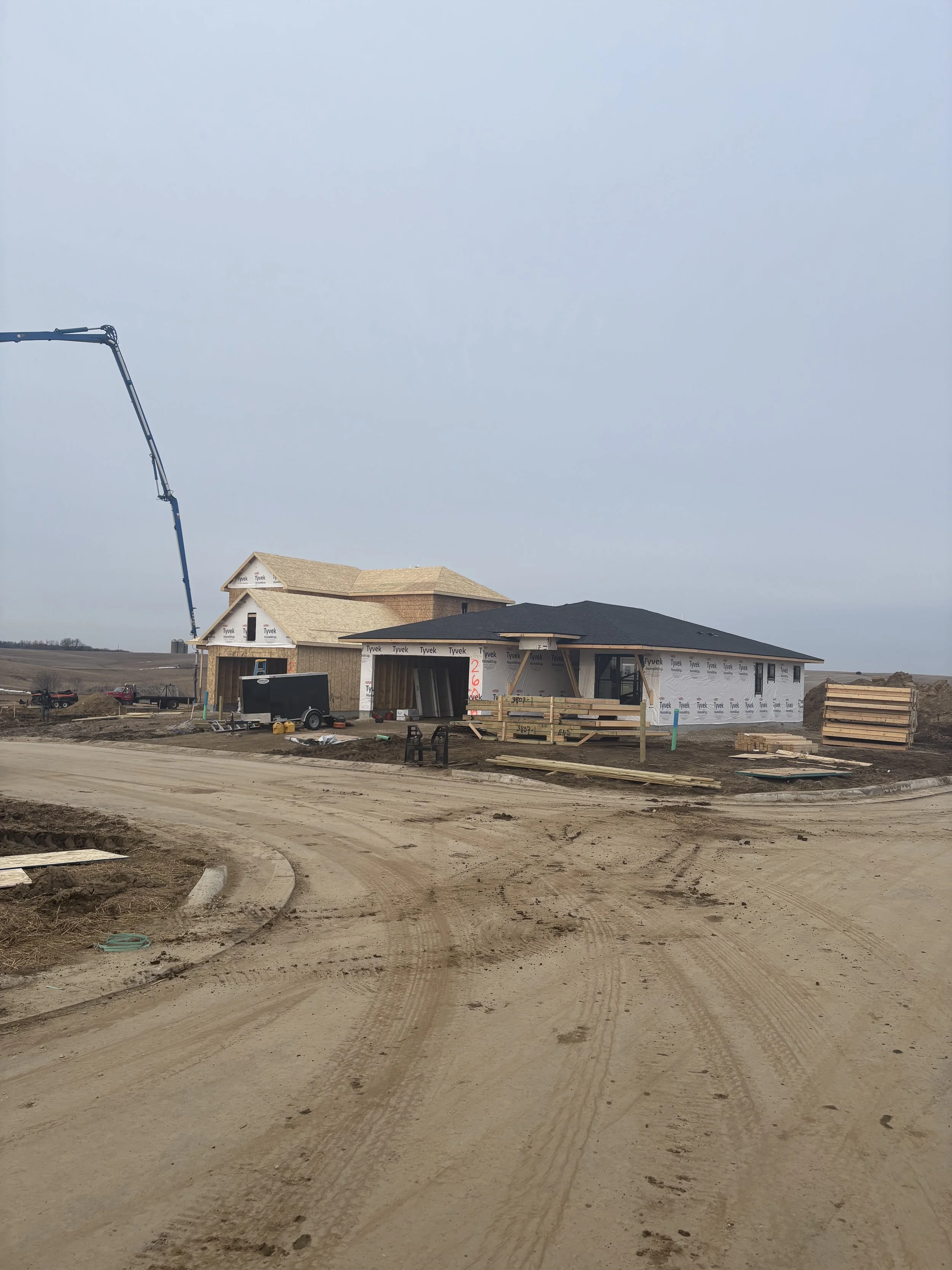 Construction site with houses under construction, showing unfinished walls and roofing, dirt road, construction equipment, and cloudy sky.