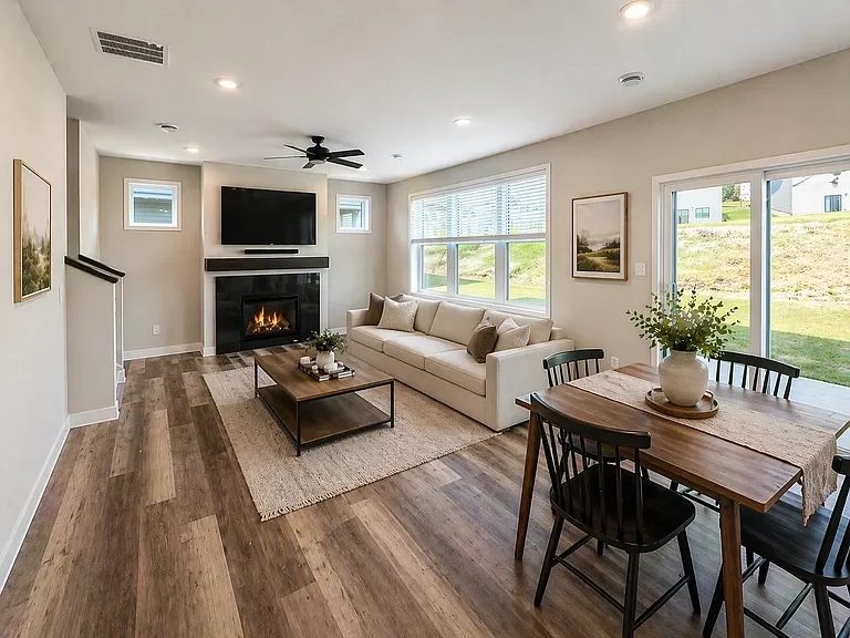 Bright living room with a white sofa, a wooden dining table with four chairs, a fireplace, a mounted TV, large windows, and artwork on the walls.