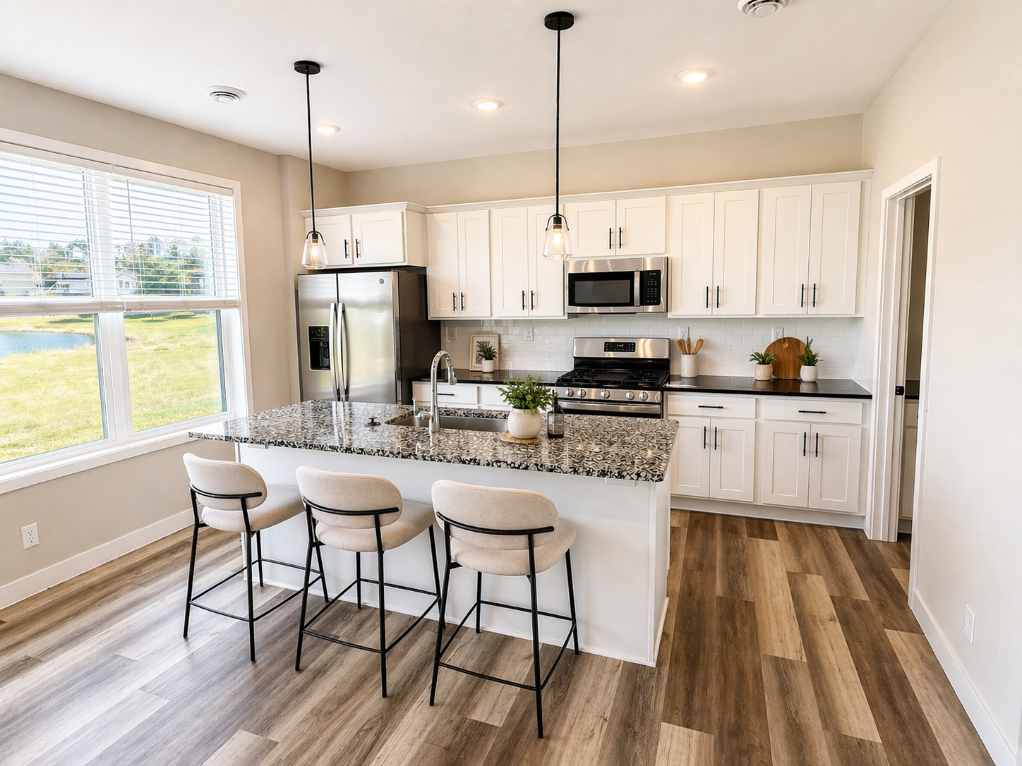 Bright modern kitchen with white cabinets, stainless steel appliances, granite island, and wood flooring, with large window showing greenery outside.