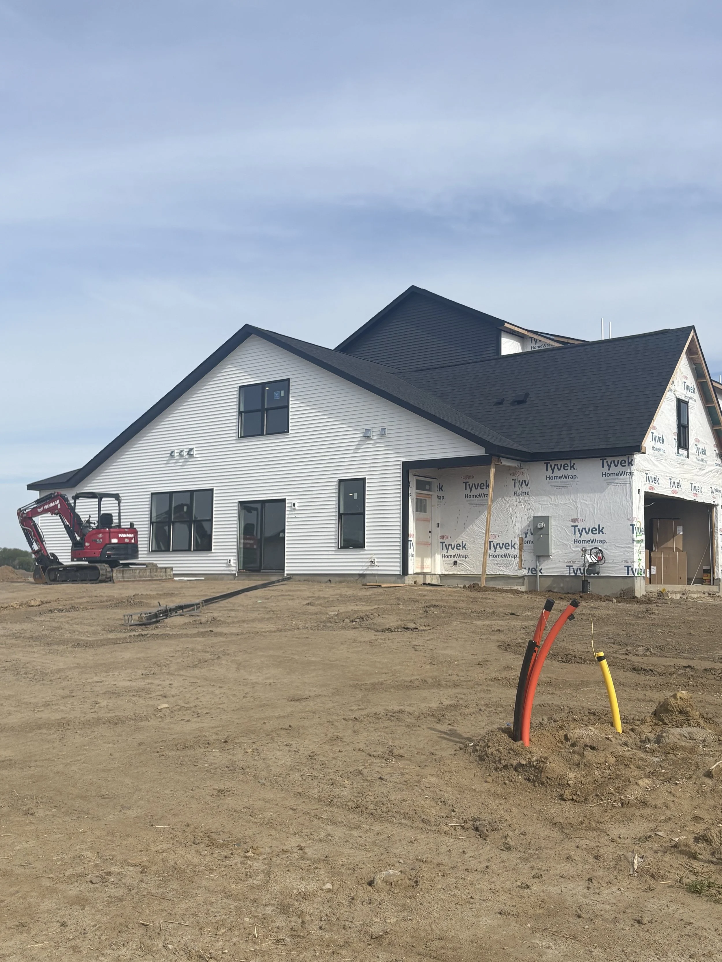 Newly built house with white siding and black roof, under construction, with exposed Tyvek wrap and construction equipment on the dirt yard.