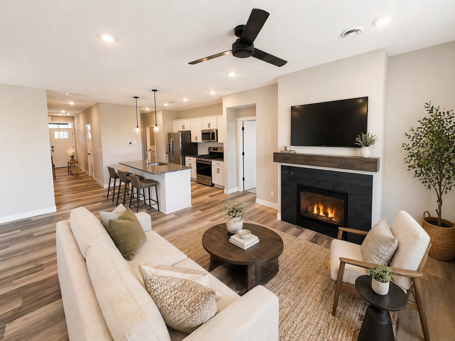Open-concept living room and kitchen with a fireplace, flat-screen TV, sofa, armchair, coffee table, and kitchen island with bar stools in a modern style with wood flooring and neutral colors.