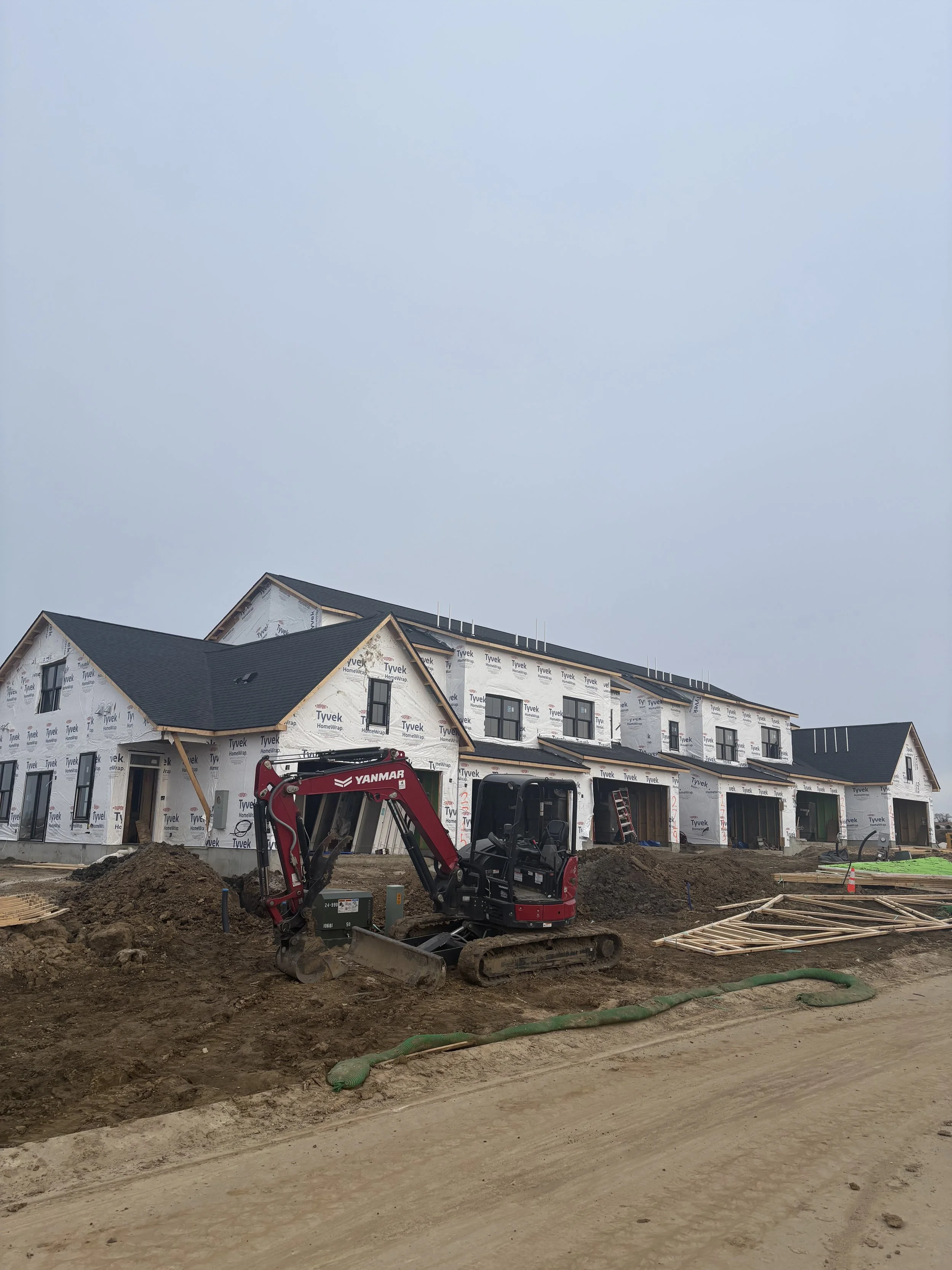 Under construction row houses with black roofs covered in Tyvek HouseWrap, construction equipment, dirt, and wooden framing materials in the foreground under a cloudy sky.