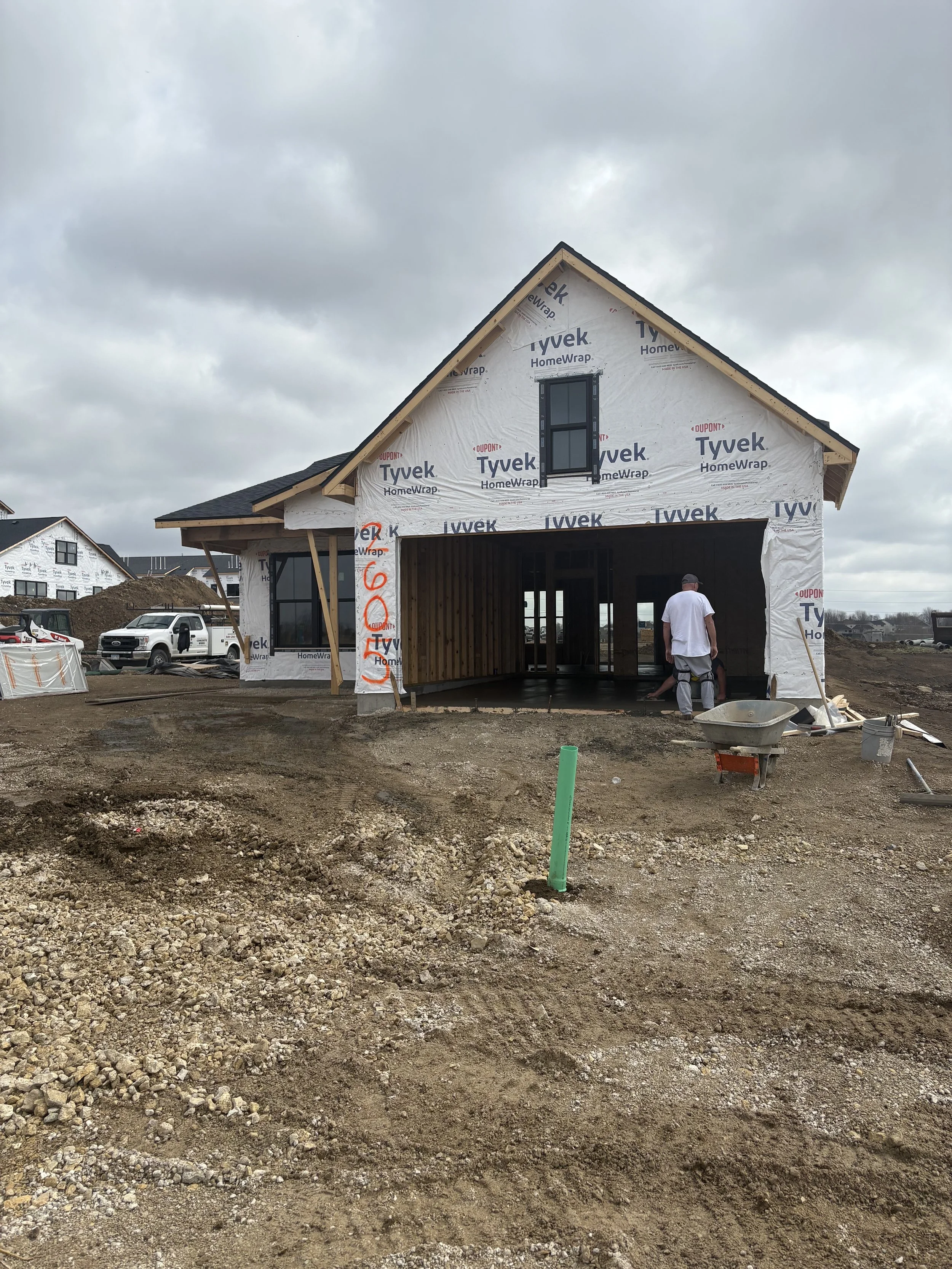 A house under construction with a partially completed wooden frame, wrapped in Tyvek HomeWrap, and with a third-floor window installed. The area surrounding the house has construction debris and soil, with some construction workers present. Overcast 