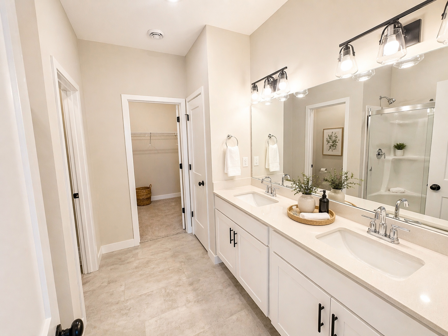 Bright bathroom with double sinks, large mirror, and walk-in shower, featuring neutral colors, white cabinetry, modern fixtures, and some decorative greenery.