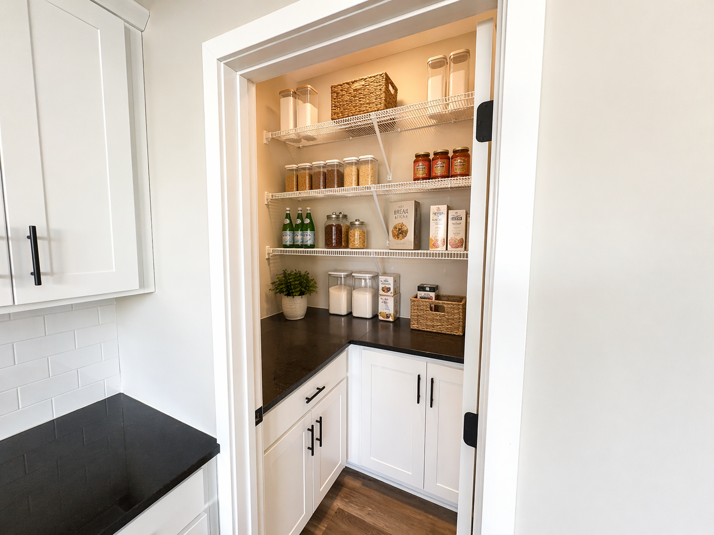 Pantry closet with white wire shelves filled with jars and boxes of food items, a small potted plant, and glass jars with flour and sugar. The pantry is inside a kitchen with white cabinets and black countertops.