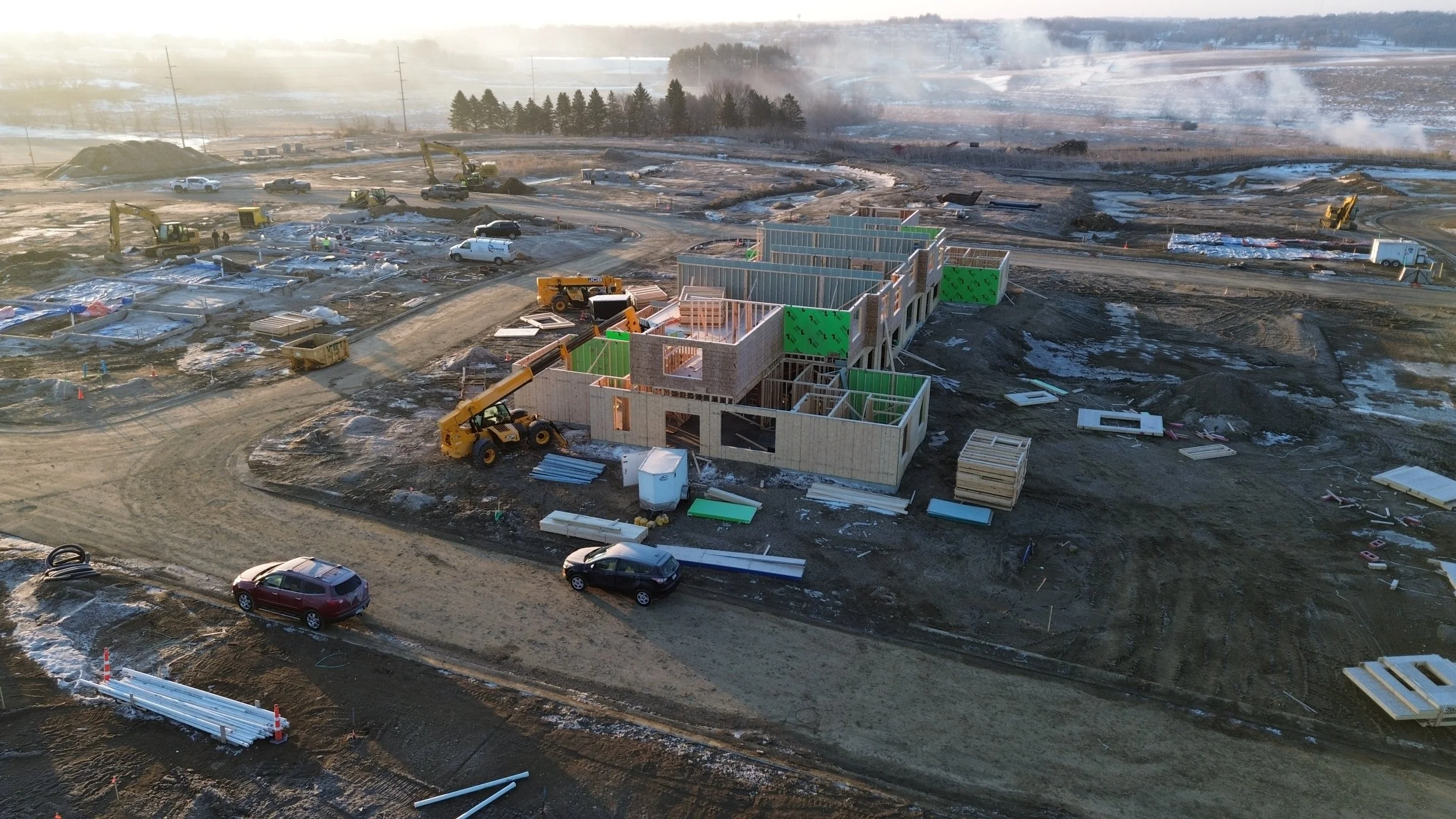 Construction site with partially built wooden frame house, construction vehicles, and scattered building materials on muddy ground, snow and steam in the background.