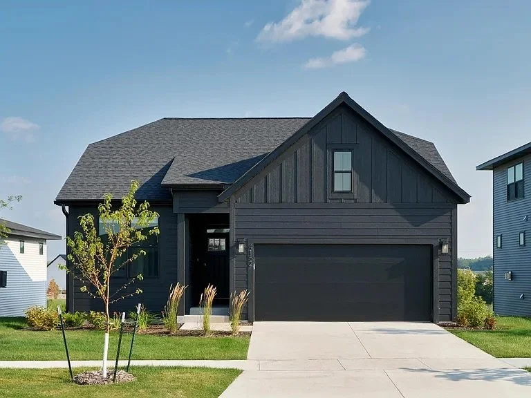A modern black two-story house with a gable roof, a small front yard with young trees and plants, and a concrete driveway in front.