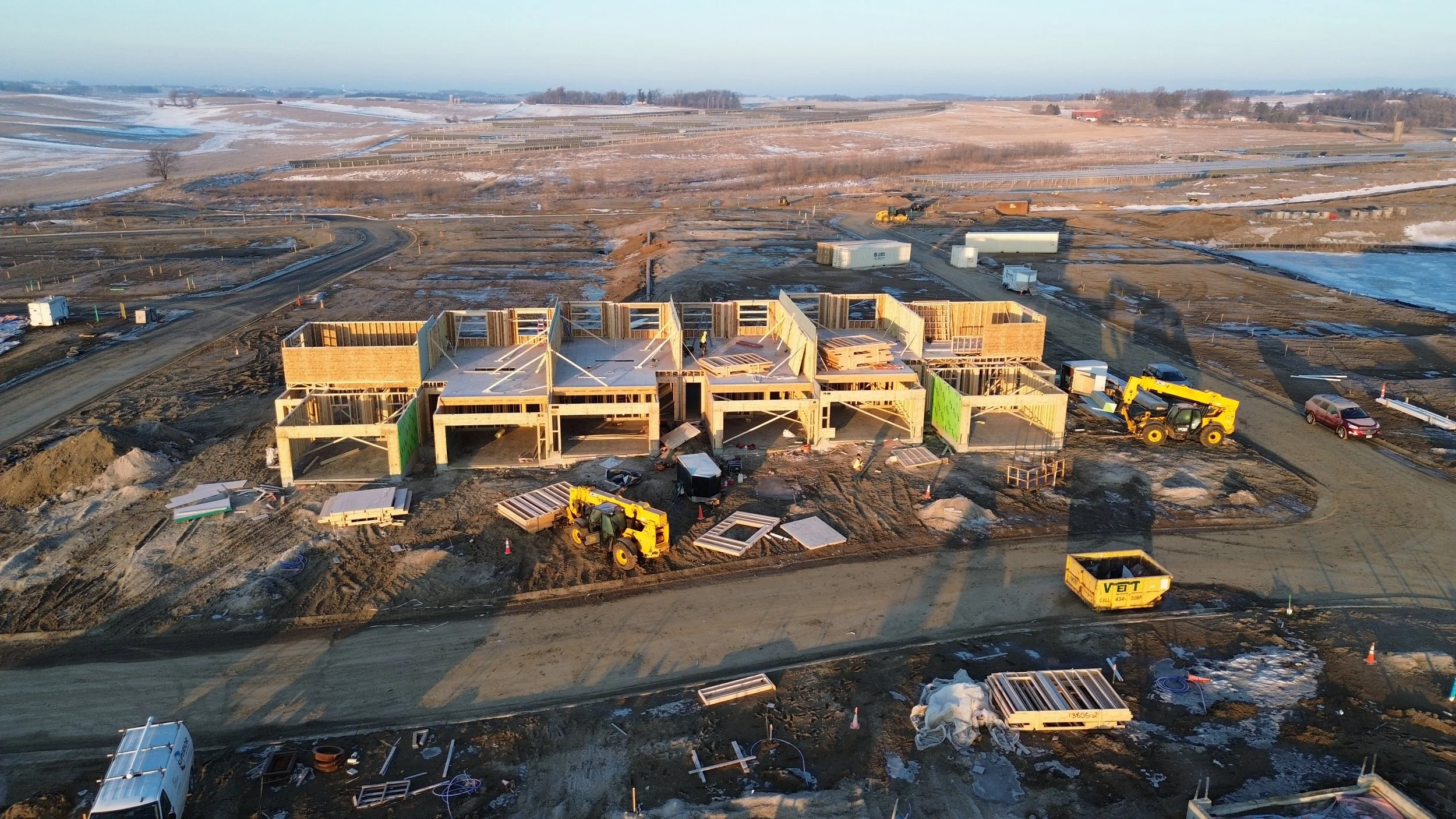 Construction site with partially built wooden structures and construction vehicles in a rural area with snow patches and open land.