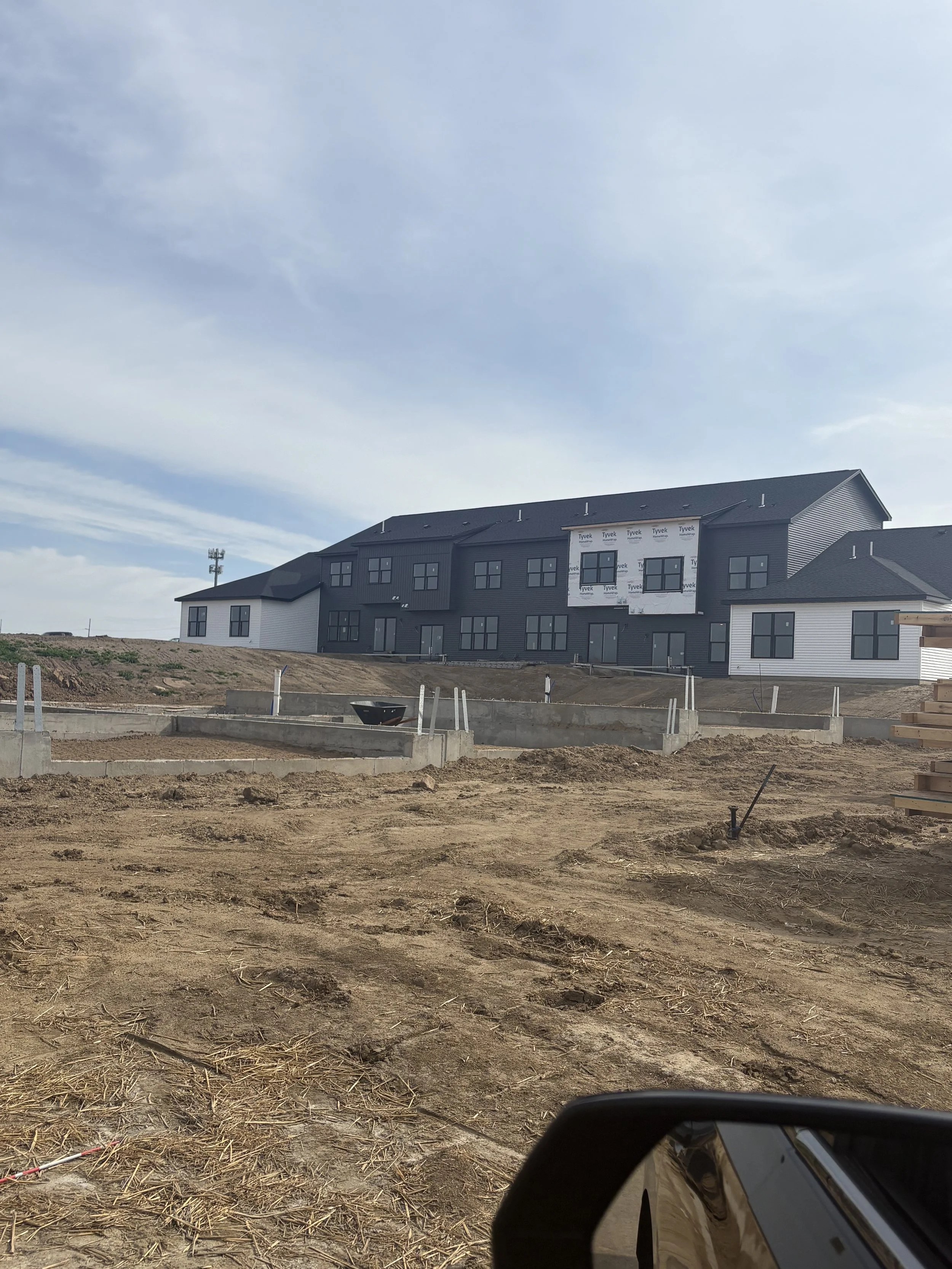 Construction site with partially built multi-story building, exposed framing, and dirt ground in the foreground.