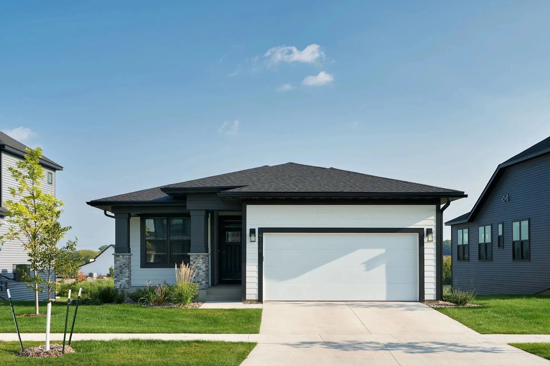 Modern two-story house with a white garage door, black front door, and gray siding. Well-maintained lawn with young trees and plants, concrete driveway, and neighboring houses with similar design under a partly cloudy sky.