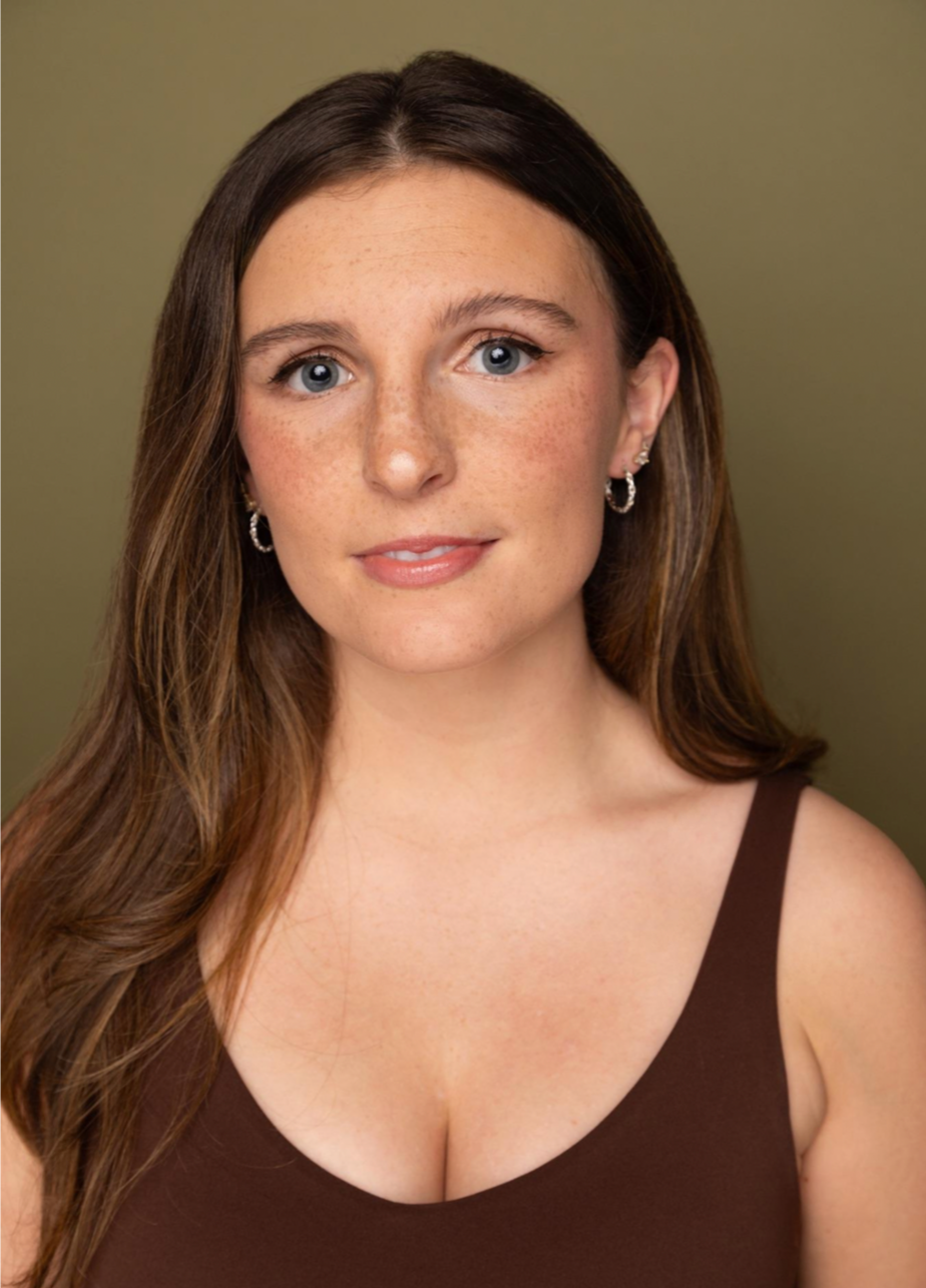 A woman with long brown hair, blue eyes, and fair skin with freckles, wearing hoop earrings and a brown top, smiling slightly against a greenish background.