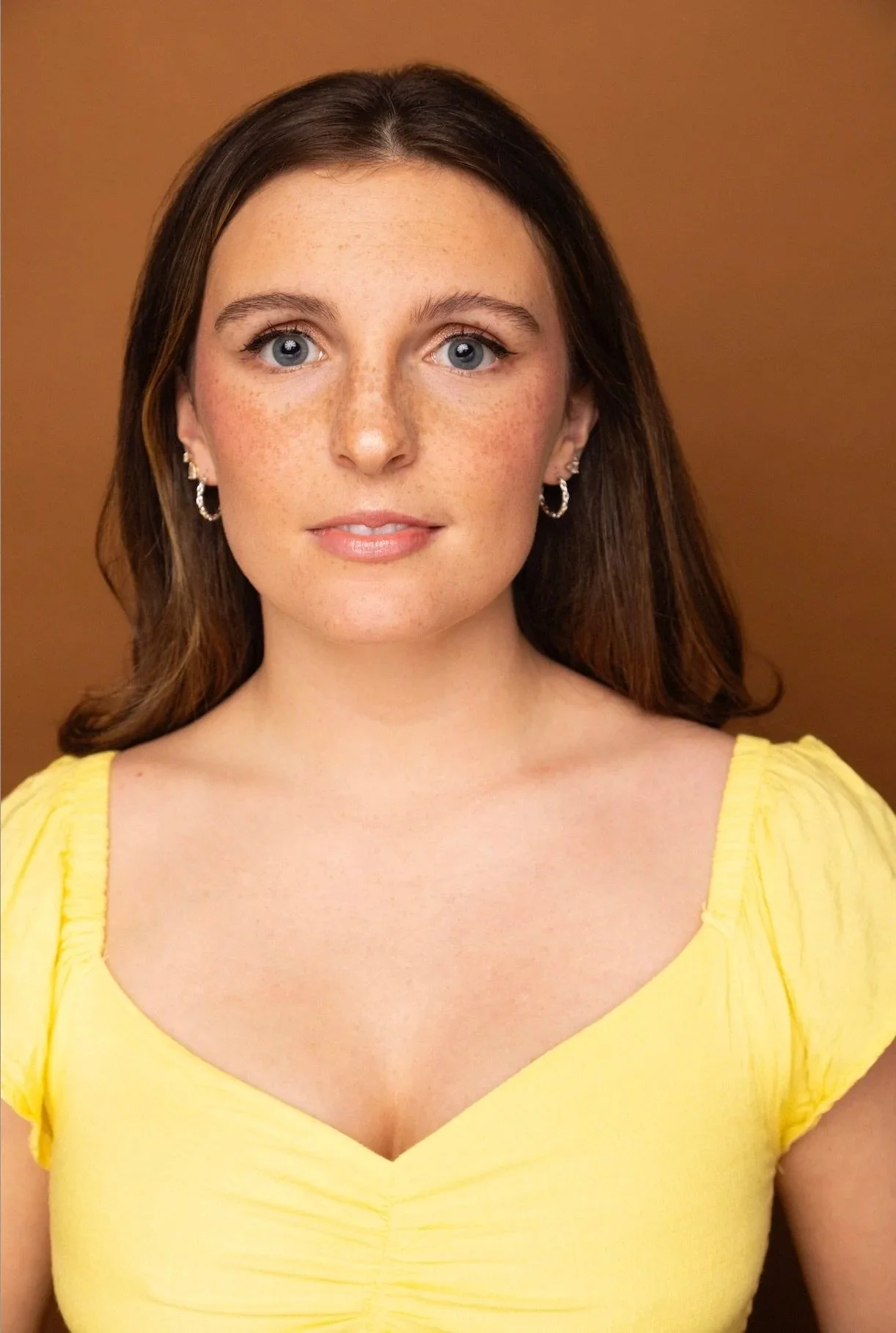 Close-up of a young woman with fair skin, blue eyes, freckles, shoulder-length brown hair, wearing earrings and a yellow top, standing against a brown background.