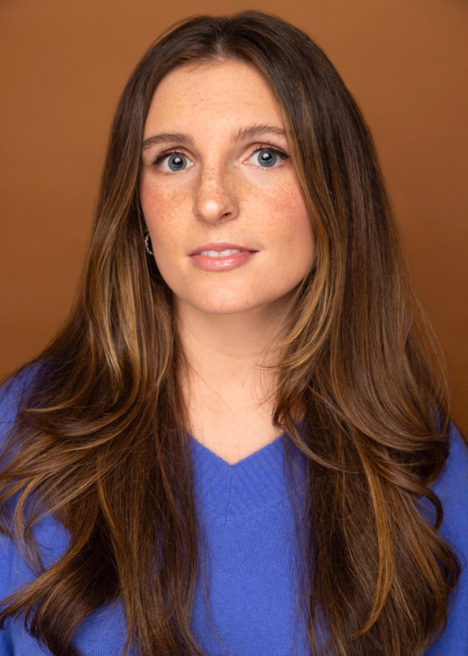 A woman with long, wavy brown hair, fair skin, blue eyes, and freckles, wearing a blue top and small earrings, against a brown background.
