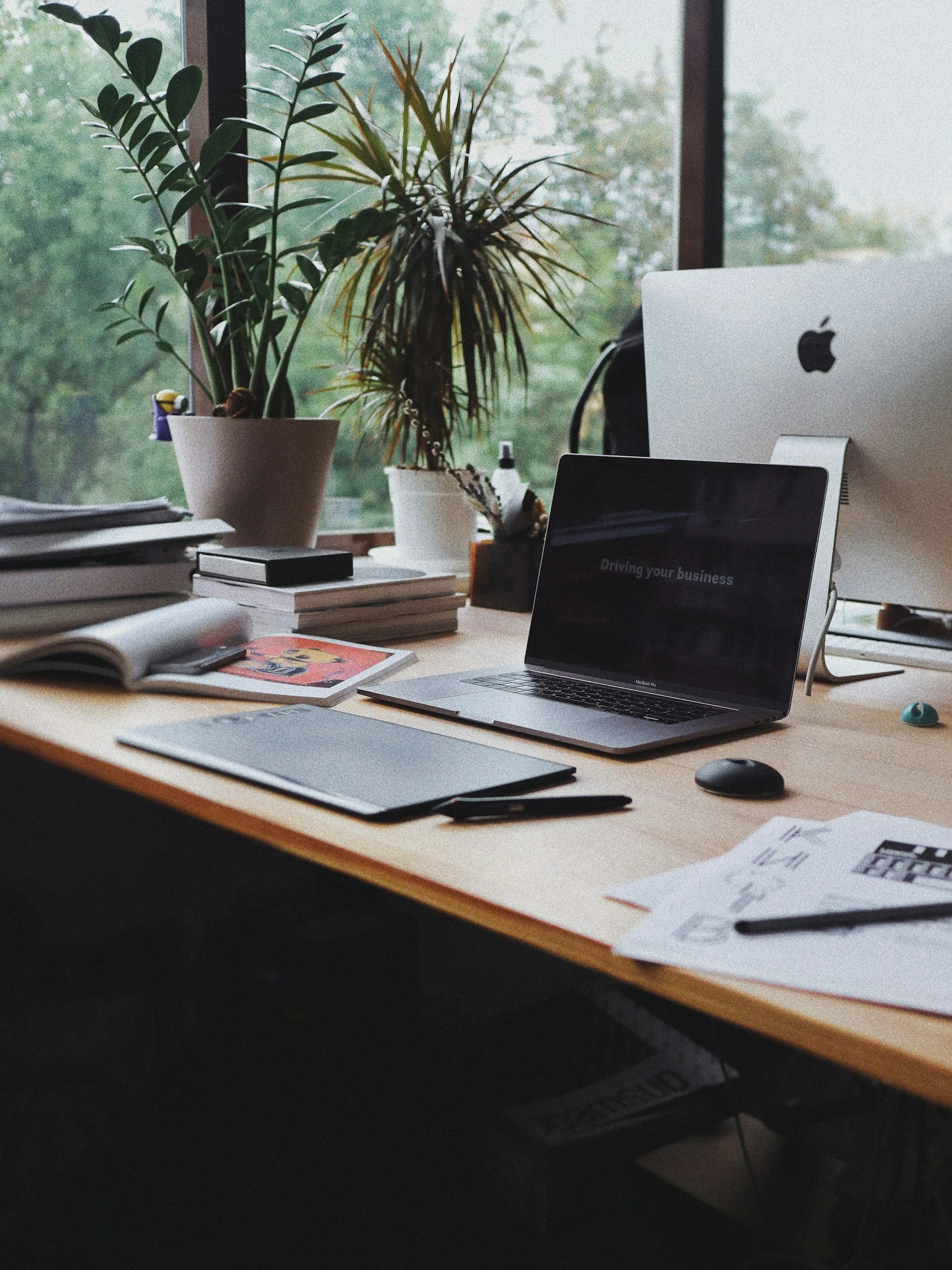 A cluttered desk near a window with two potted plants, a MacBook Pro laptop, a mouse, papers, magazines, a pen, and a laptop stand.