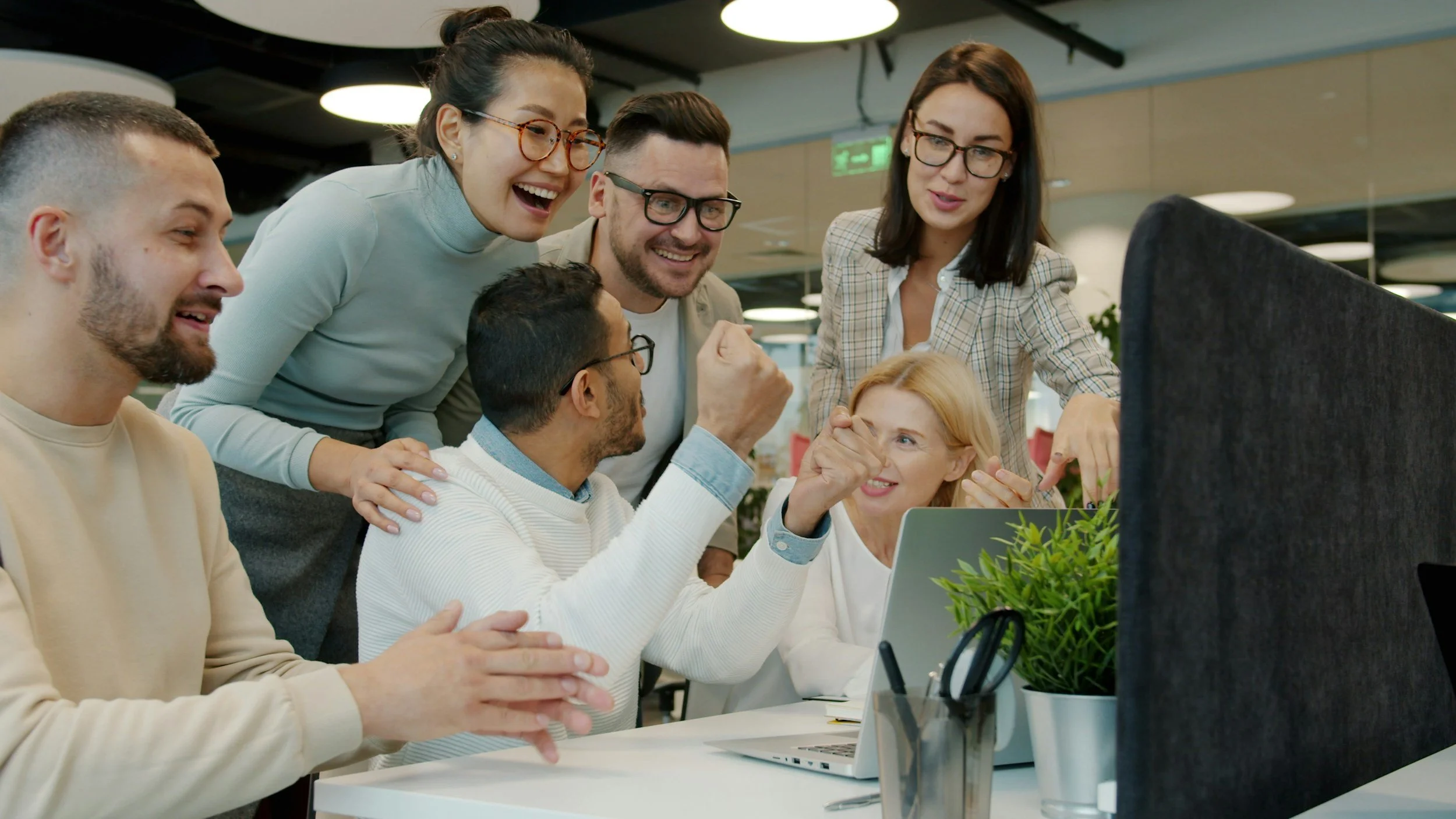 A diverse group of six coworkers gathered around a computer, smiling, laughing, and engaging enthusiastically in a collaborative office space.